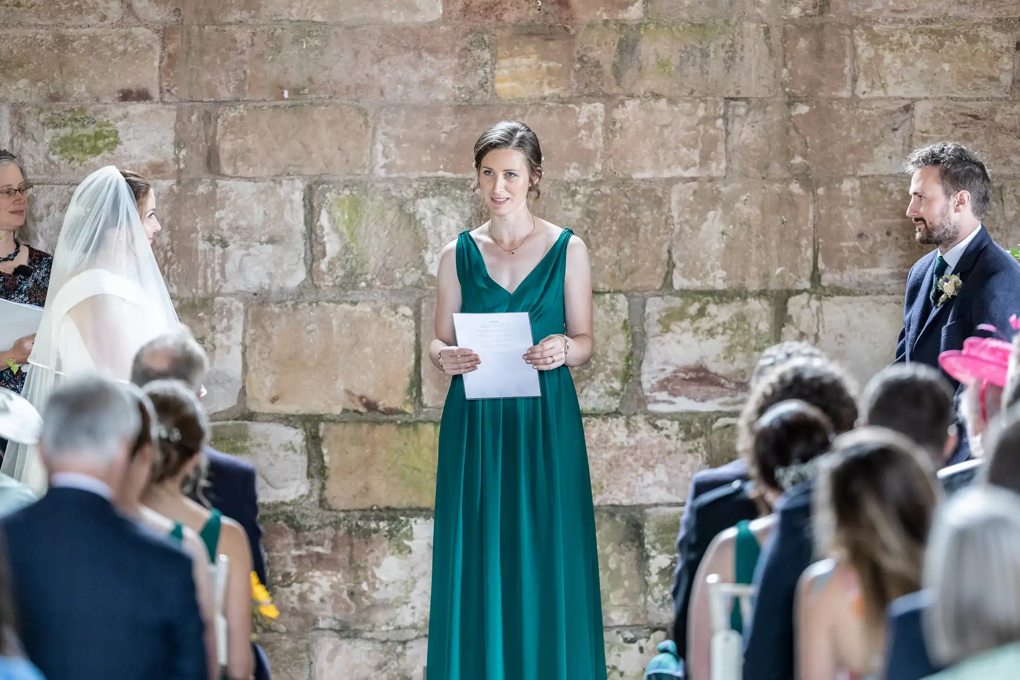 A woman in a green dress speaks at a wedding ceremony, holding a piece of paper. The bride and groom stand to the sides, and guests are seated facing them.