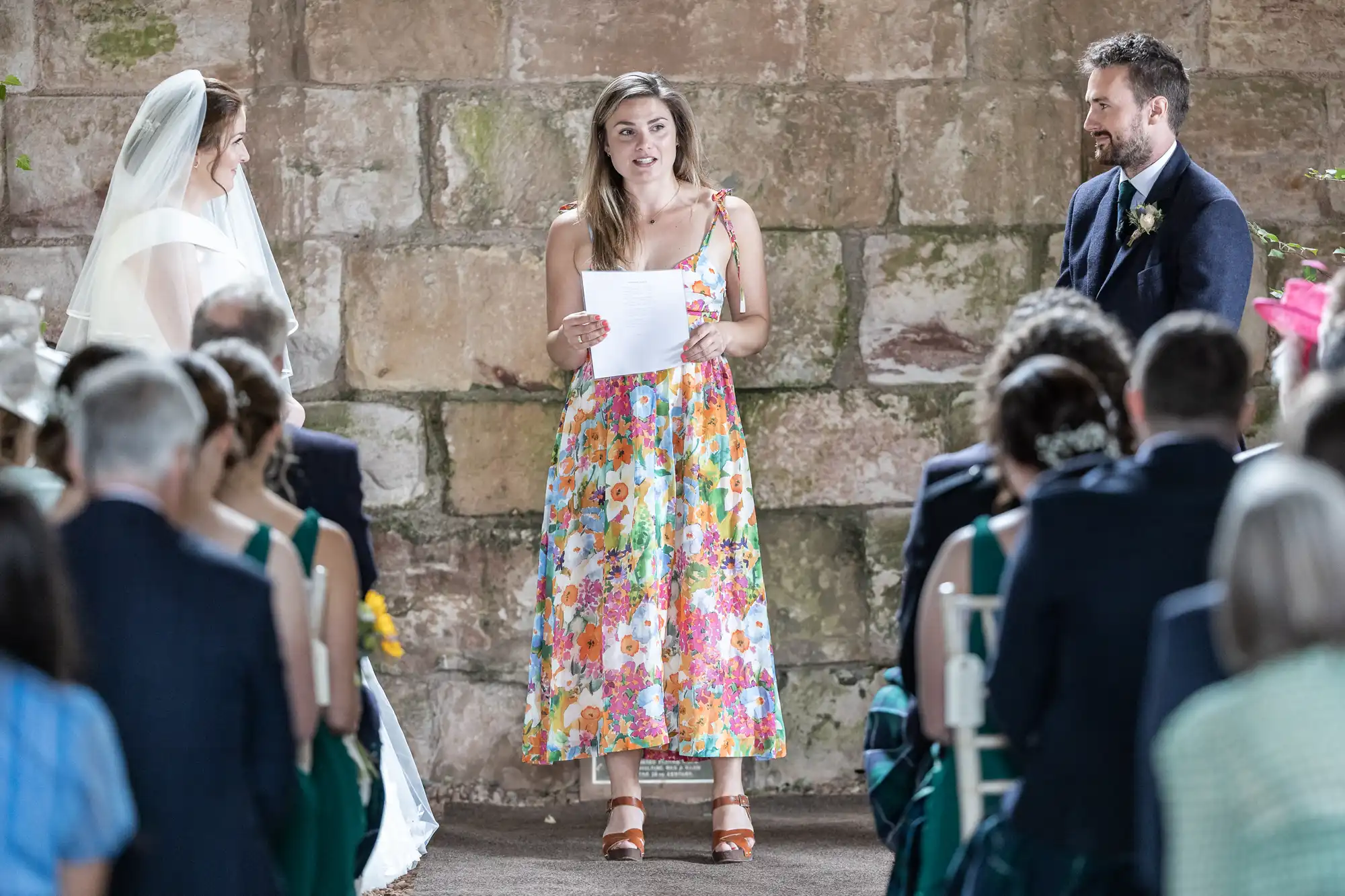 A woman in a floral dress stands and reads from a paper at a wedding ceremony, with a bride and groom facing her and guests seated in the foreground.