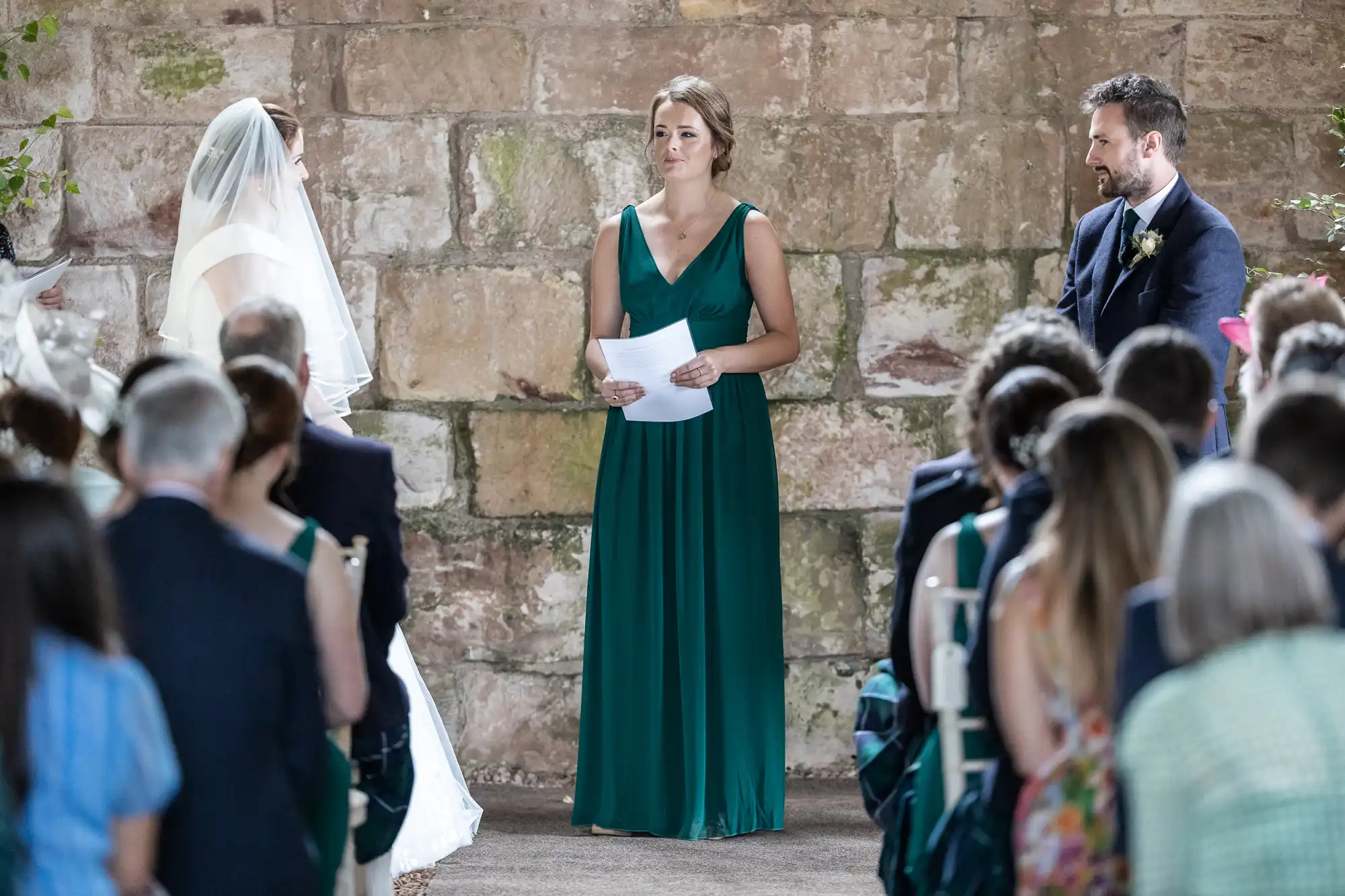 A woman in a green dress stands holding papers between a bride and groom during a wedding ceremony. Guests are seated and facing the couple.