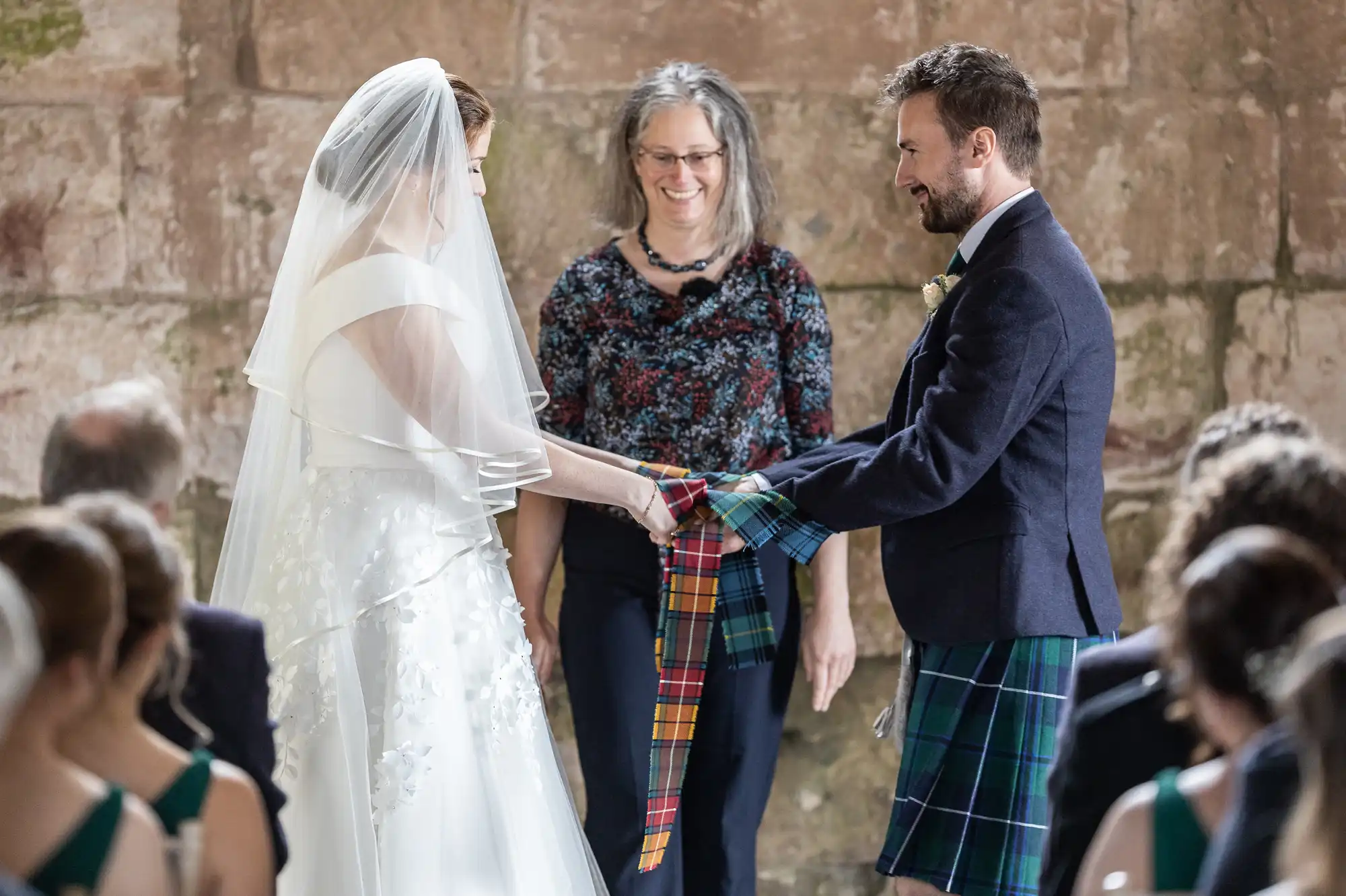 A bride and groom hold hands with a tartan cloth wrapped around them during a wedding ceremony, with an officiant standing behind them.