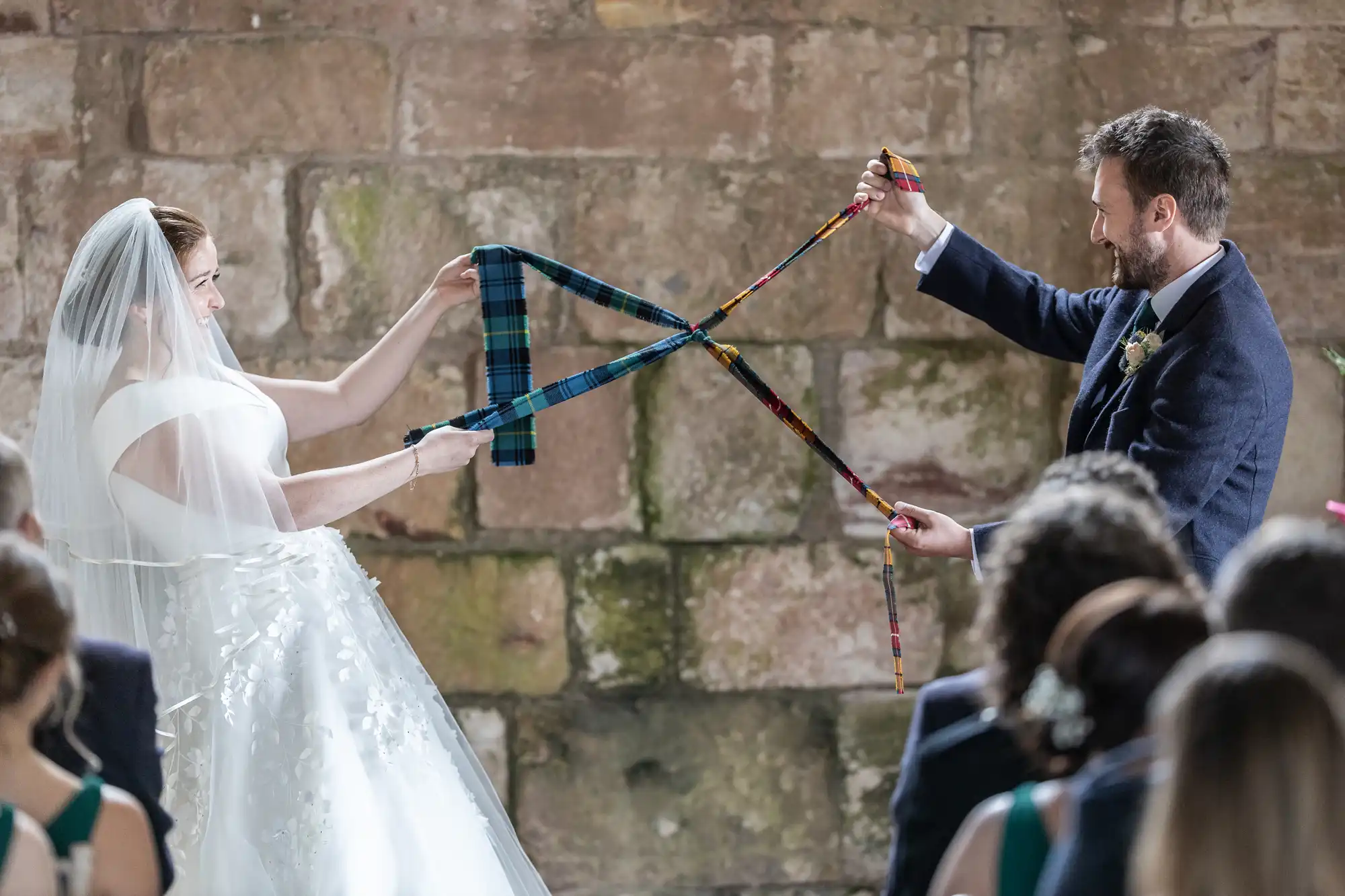 A bride and groom, dressed in wedding attire, are holding and intertwining tartan ribbons in front of an audience during a ceremony. A stone wall is visible in the background.