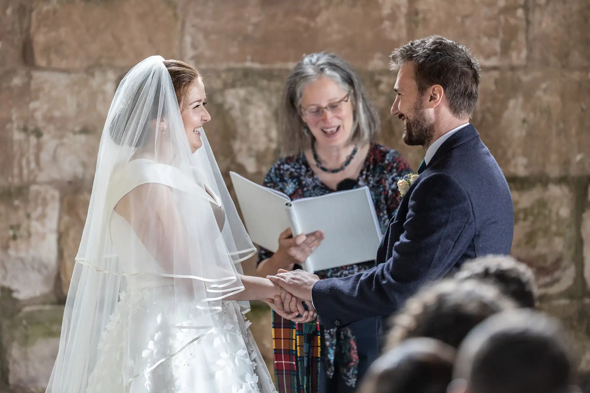 A bride and groom stand facing each other, holding hands, and smiling during their wedding ceremony officiated by a person holding an open book.