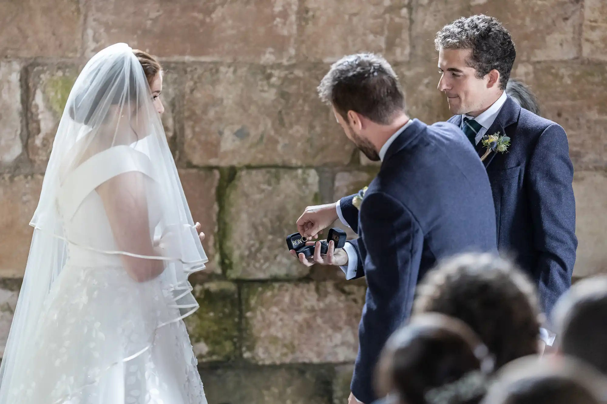 A bride and groom stand facing each other in a stone-walled venue, while a man in a dark suit hands them small black boxes, possibly containing wedding rings.