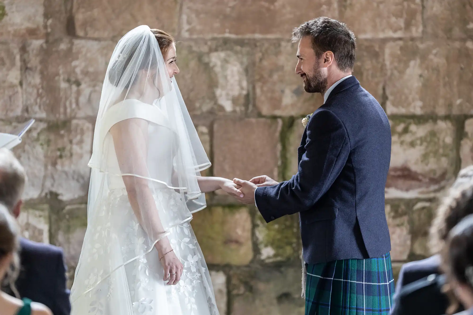 A bride and groom hold hands and exchange rings during their wedding ceremony in front of a stone wall background.