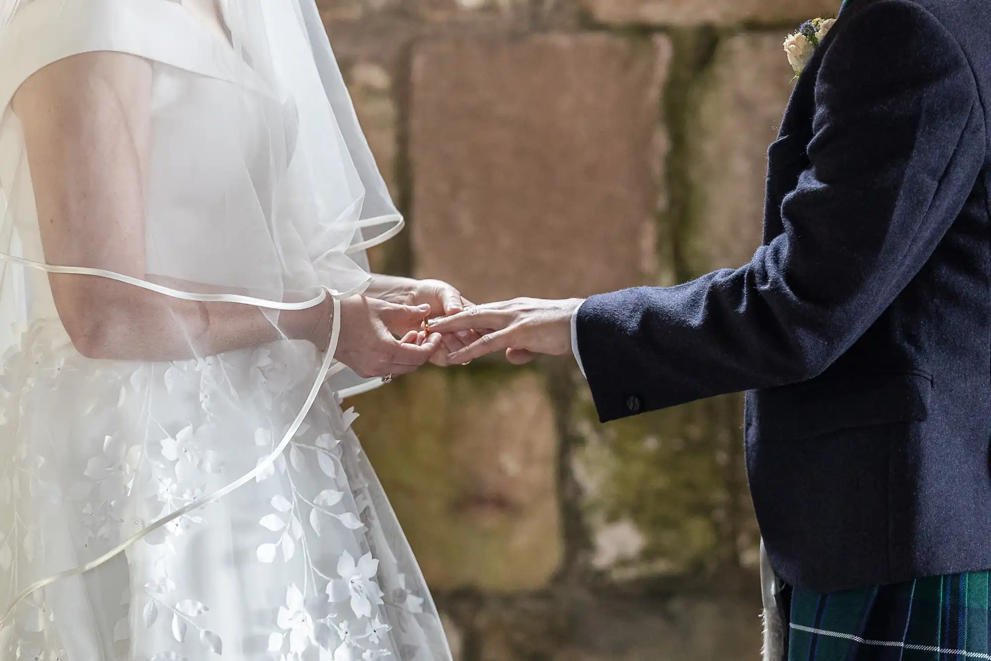 A bride and groom exchange rings during their wedding ceremony, with the bride's white dress and veil and the groom's dark suit visible.