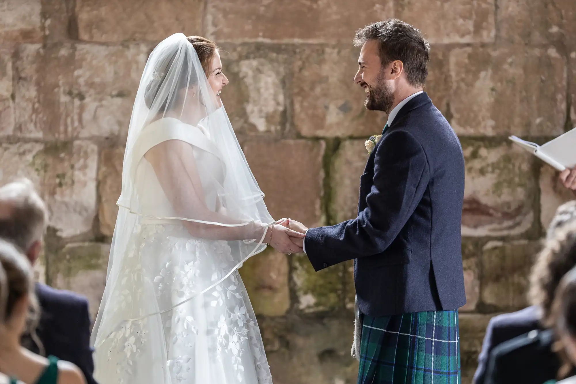 A bride and groom, dressed in wedding attire, stand facing each other holding hands, smiling, during their wedding ceremony in a rustic setting.