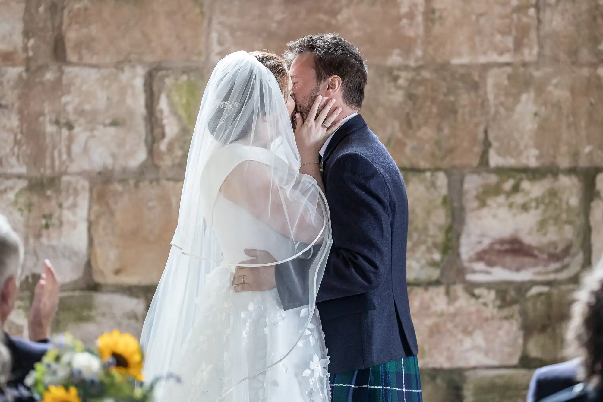 A couple in wedding attire shares a kiss, with the bride in a veil and the groom in a dark suit jacket and tartan kilt, standing against a stone wall backdrop.