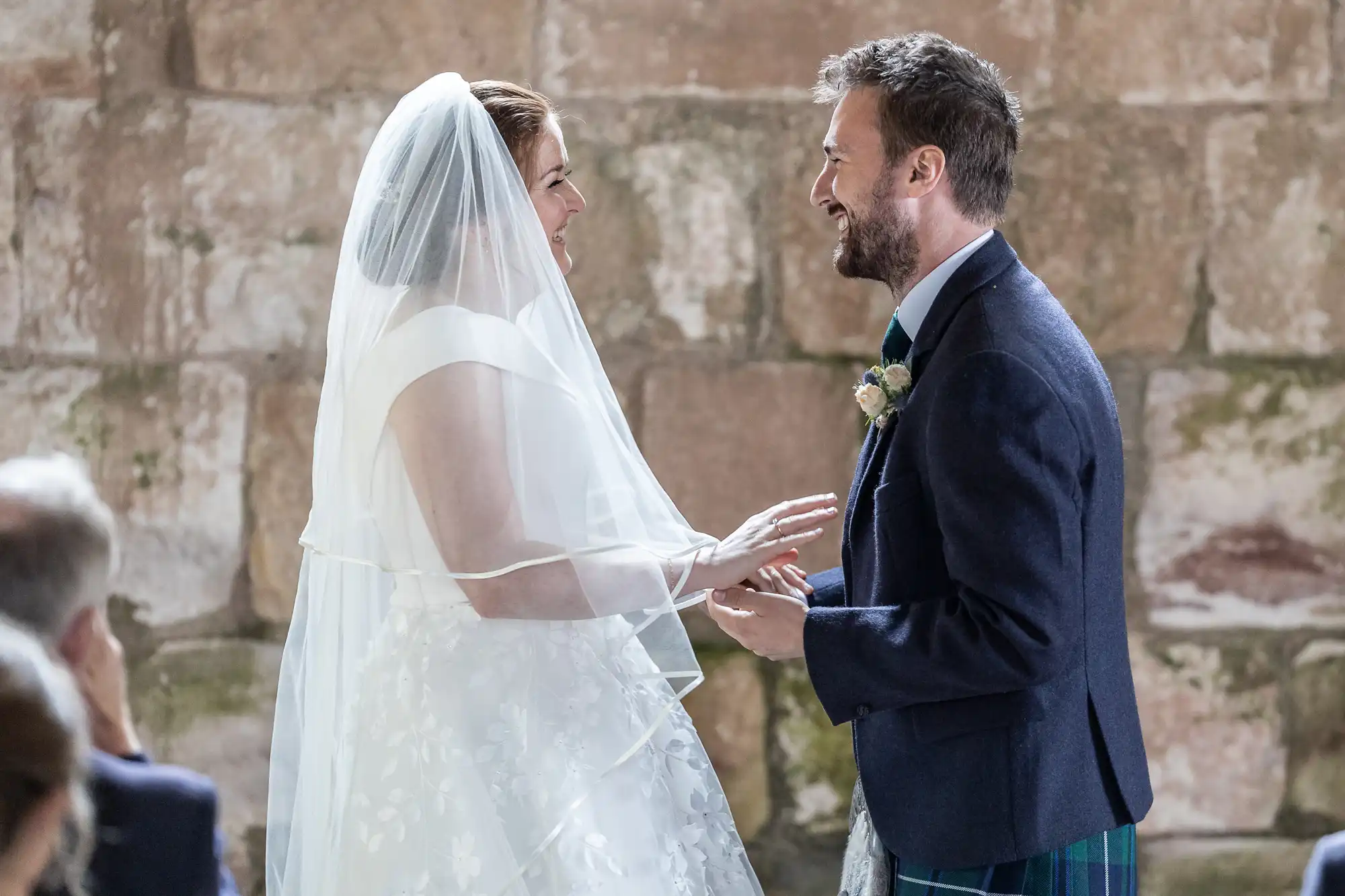 A bride and groom exchange vows in front of a stone wall, both smiling and holding hands. The bride wears a veil and white dress, while the groom is in a suit and kilt.