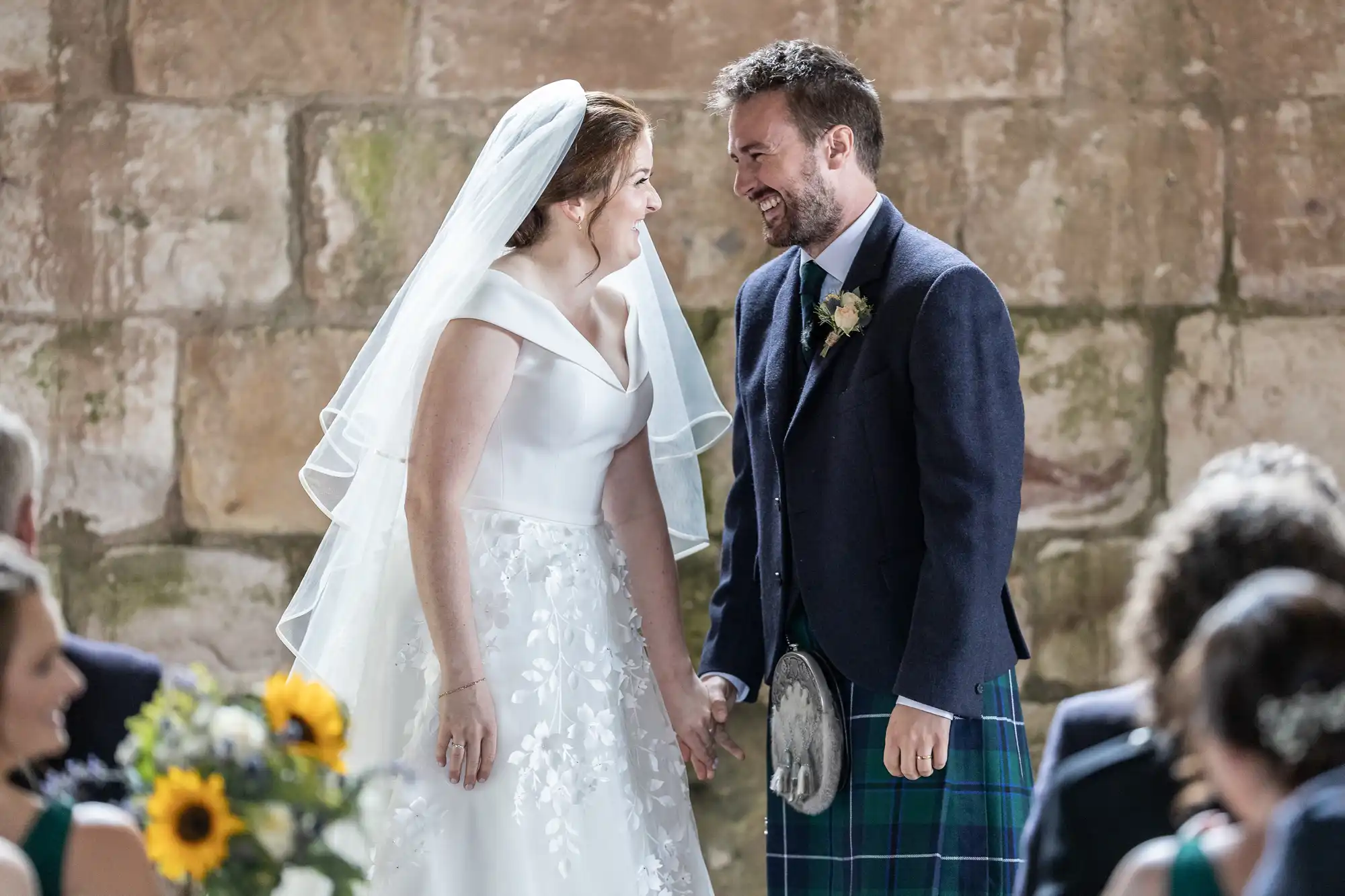 A bride in a white dress and veil, and a groom in a kilt, hold hands and smile at each other during their wedding ceremony in front of a stone wall.