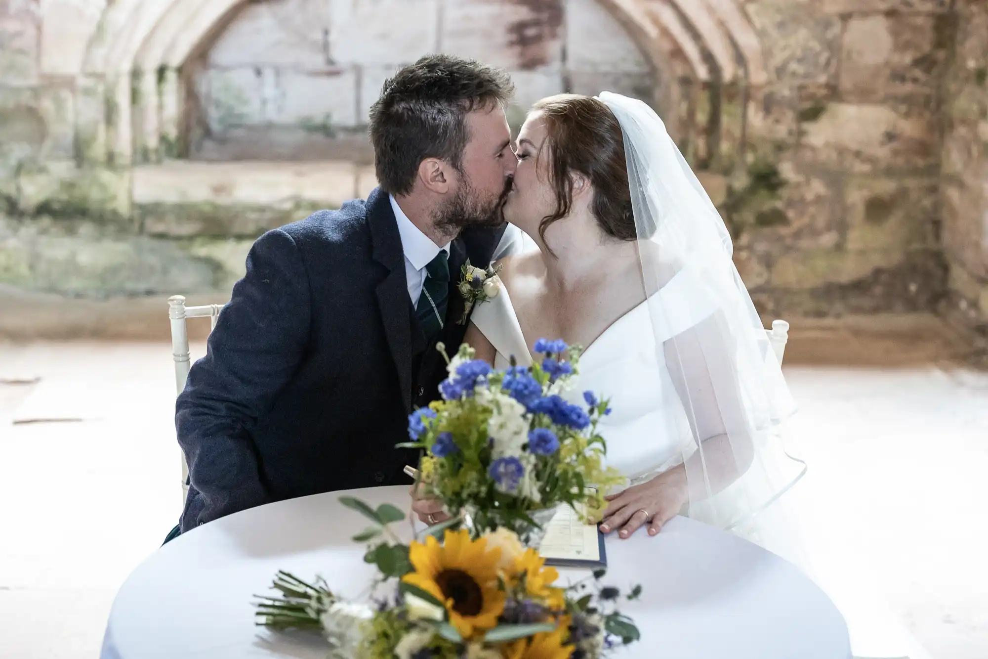 A bride and groom share a kiss while seated at a table with a bouquet of flowers during their wedding ceremony.