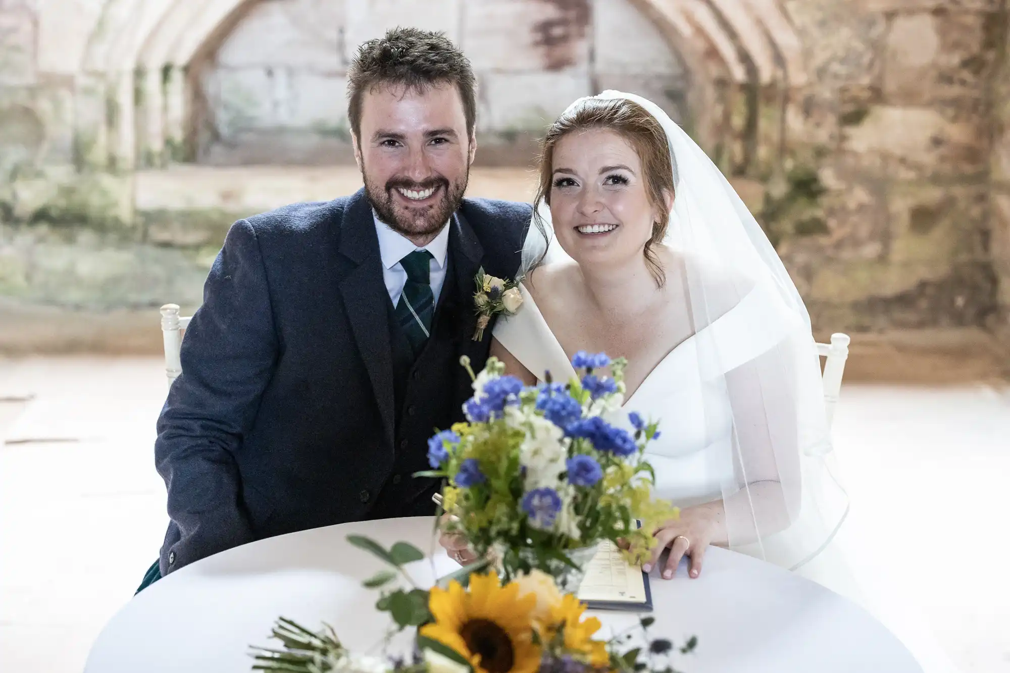 A smiling couple dressed in wedding attire sits at a table with a floral bouquet in front of them. The background features rustic stone walls and an arched doorway.