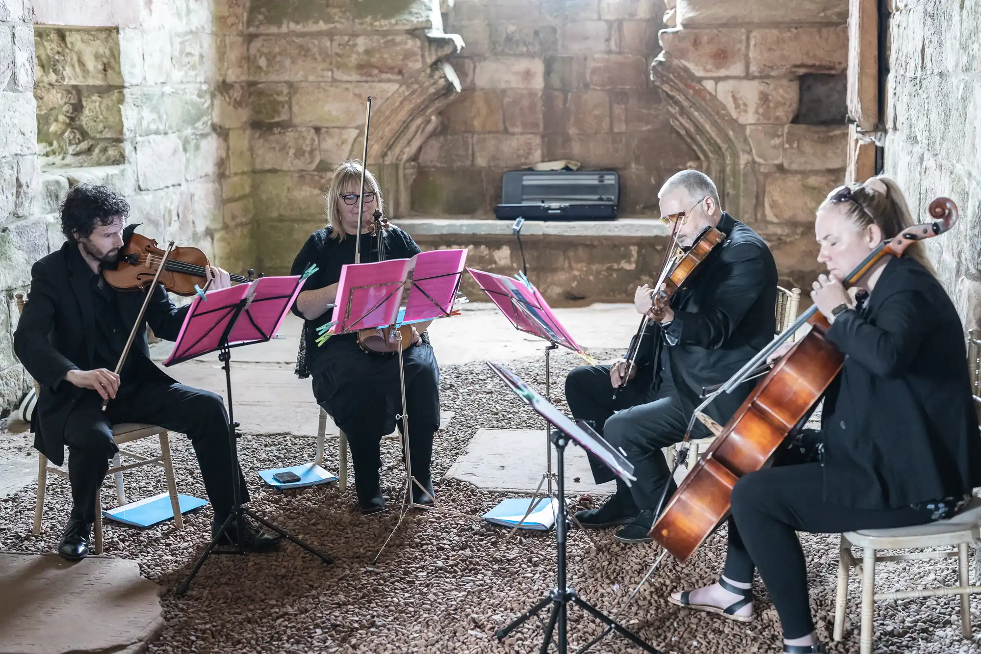 A string quartet, consisting of two violinists, a violist, and a cellist, perform in a rustic, stone-walled setting on a gravel floor. Red music stands hold their sheet music.