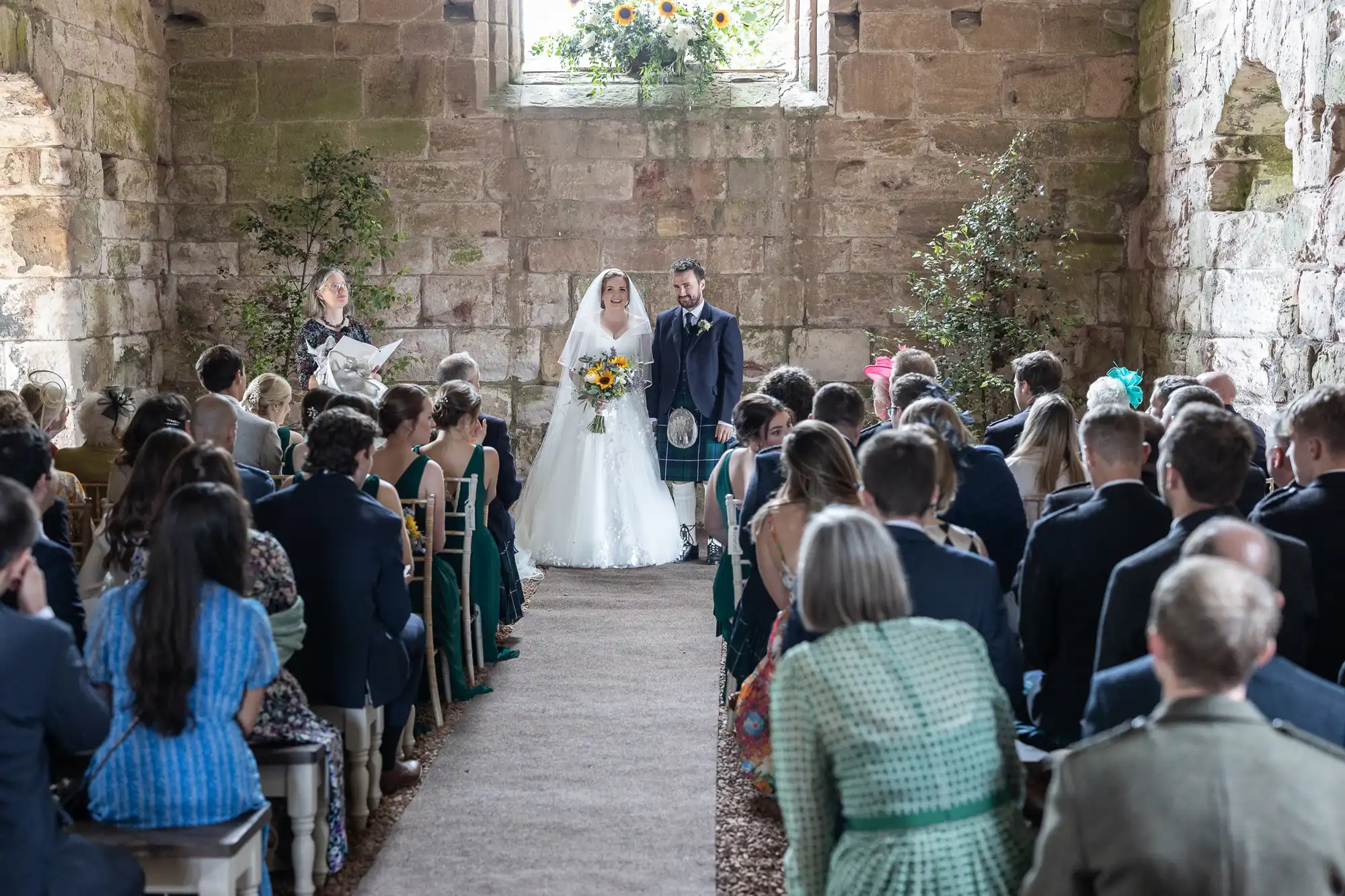 A bride and groom stand at the altar during their wedding ceremony in an old stone building, with guests seated on both sides. The bride holds a bouquet of sunflowers.