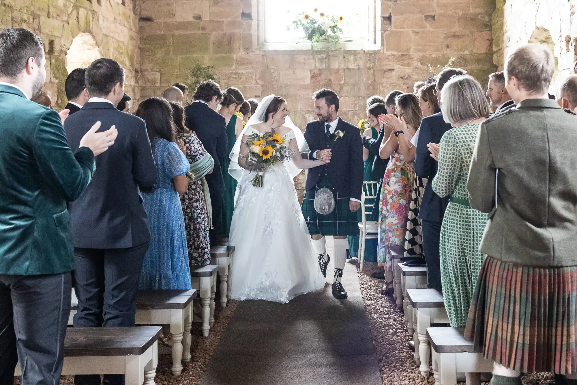 A bride and groom walk down the aisle of a rustic church, smiling and holding hands, while guests stand and applaud.