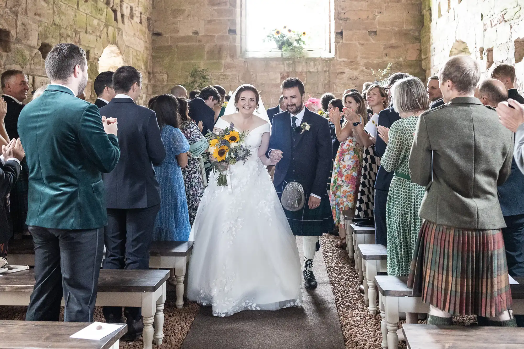 Bride and groom walk down the aisle of a rustic, stone-walled chapel, holding hands. The bride holds a sunflower bouquet while the groom wears a kilt. Guests stand and applaud on either side.