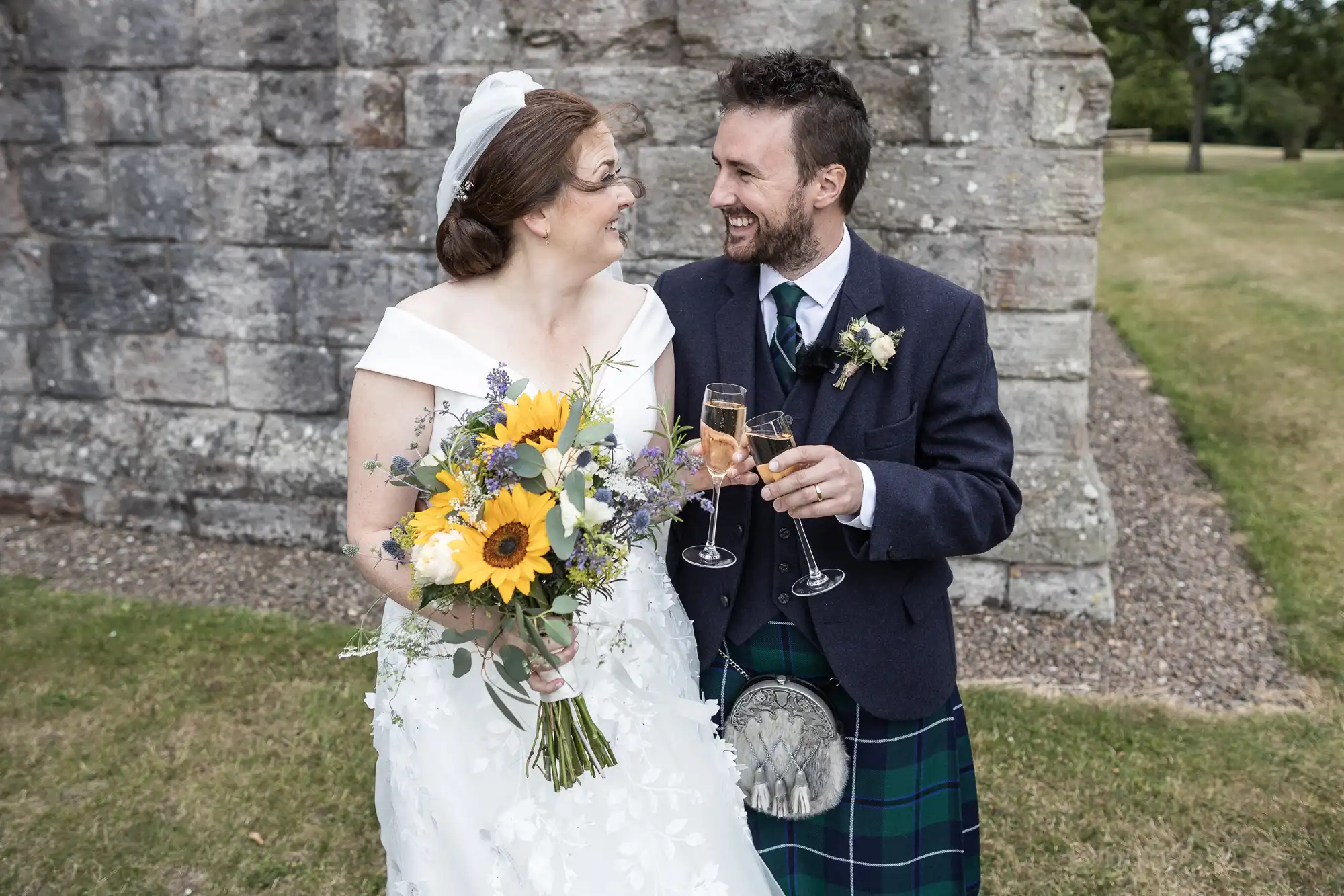 A bride in a white gown holding a bouquet and a groom in a kilt share a joyful moment with champagne glasses in hand, standing in front of a stone wall outdoors.