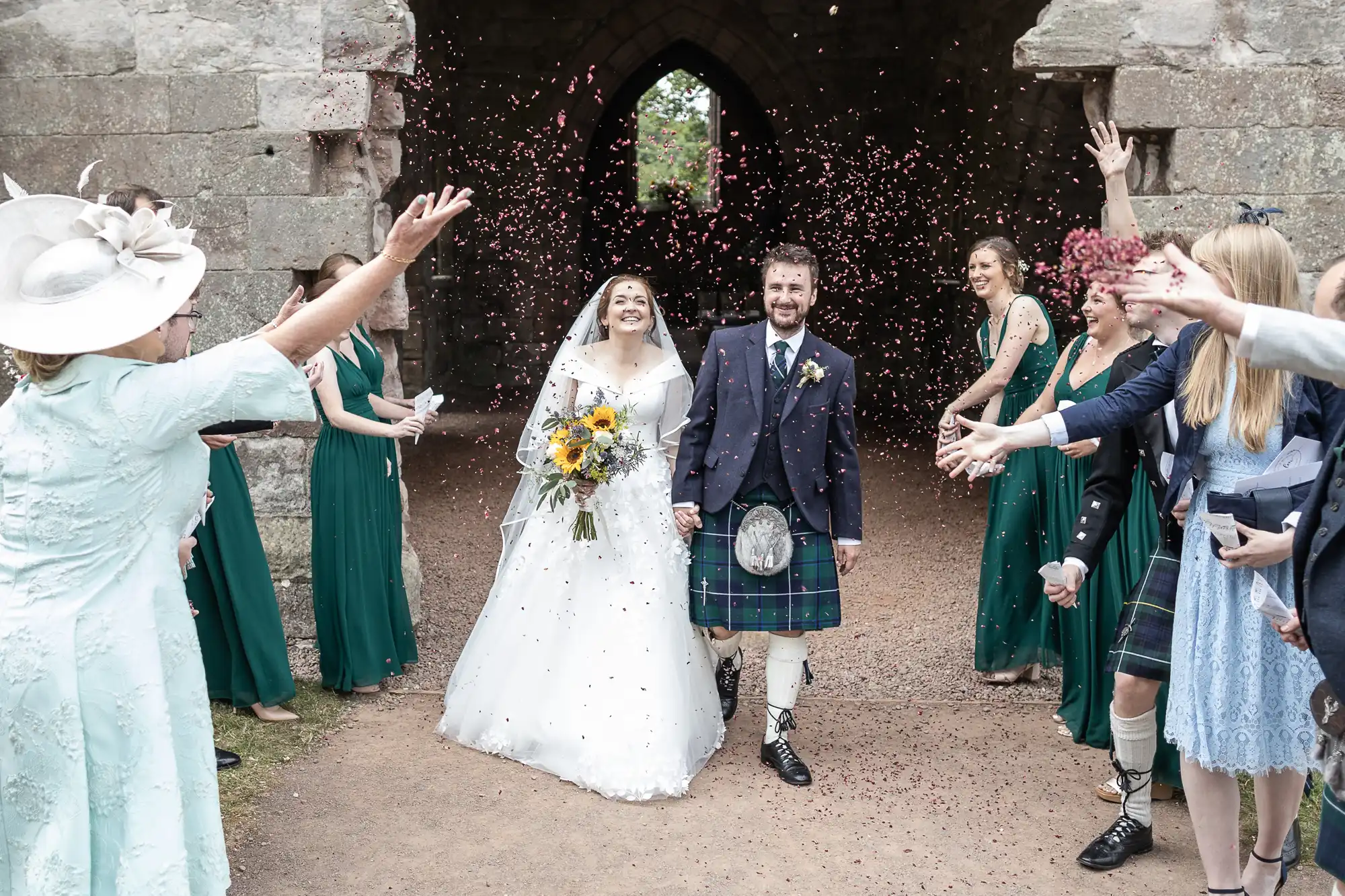 A newlywed couple in wedding attire walks under an archway while guests throw confetti. The bride holds a sunflower bouquet, and the groom wears a kilt. Bridesmaids in green dresses stand nearby.