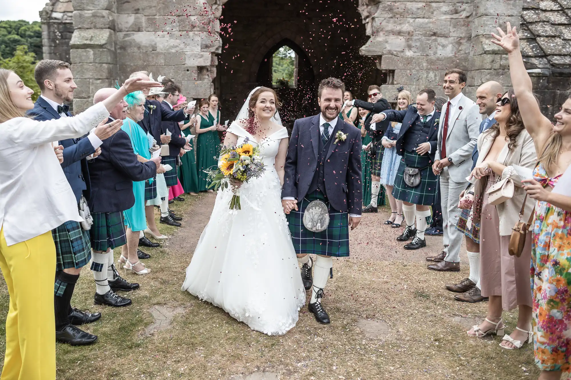 A bride and groom walk hand in hand under a shower of confetti, surrounded by friends and family, some wearing kilts, outside a stone archway.