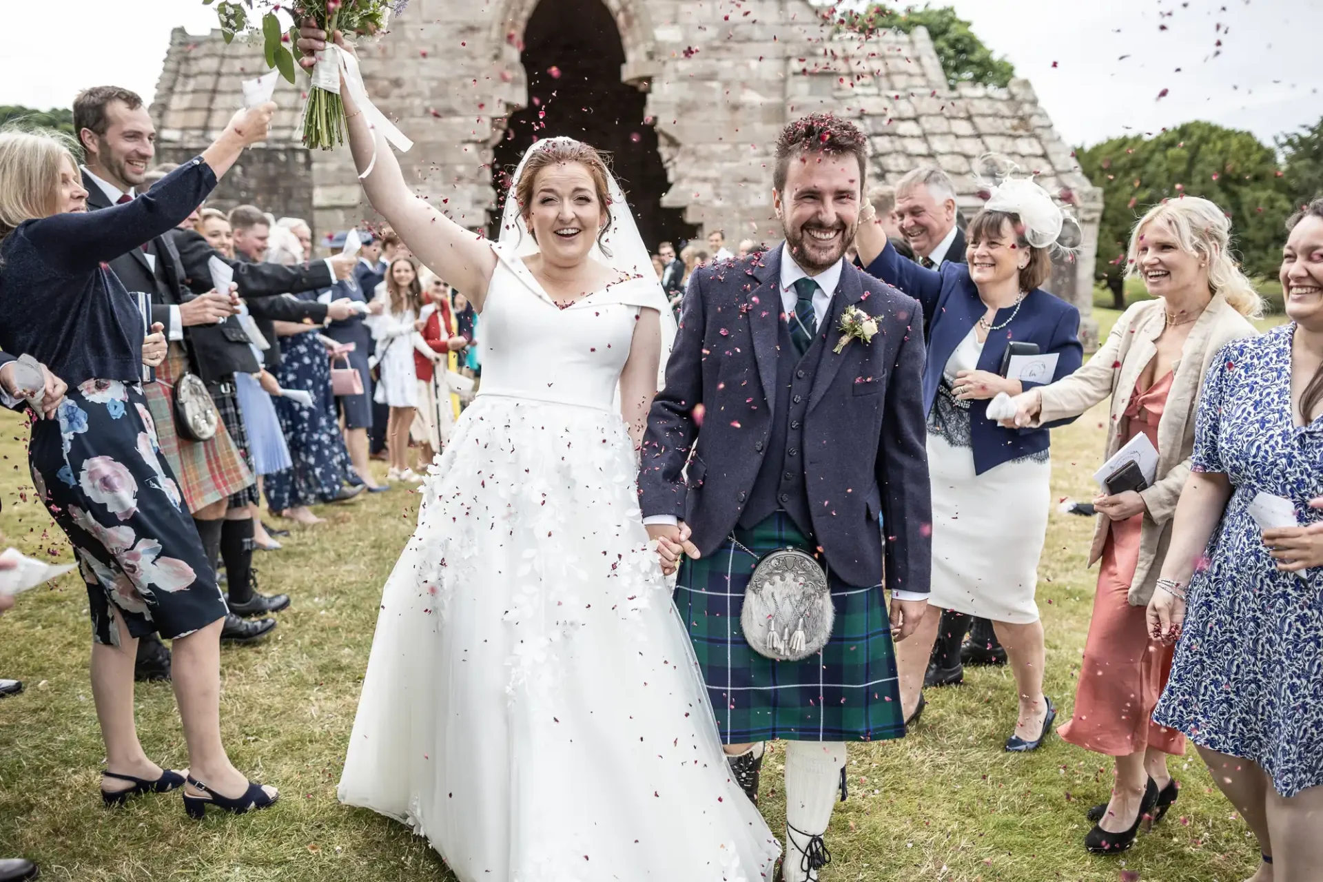 Confetti wedding photo: A bride and groom walk hand in hand smiling while wedding guests throw confetti. The groom is wearing a kilt and the bride a white gown, with an arched stone structure in the background.