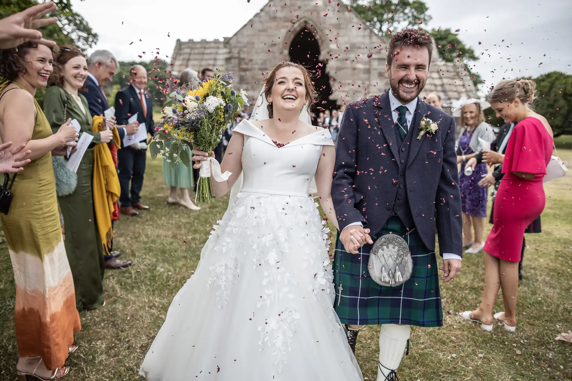 A bride in a white dress and a groom in a kilt walk joyfully down an outdoor aisle as guests clapping throw flower petals at them in front of a stone building.