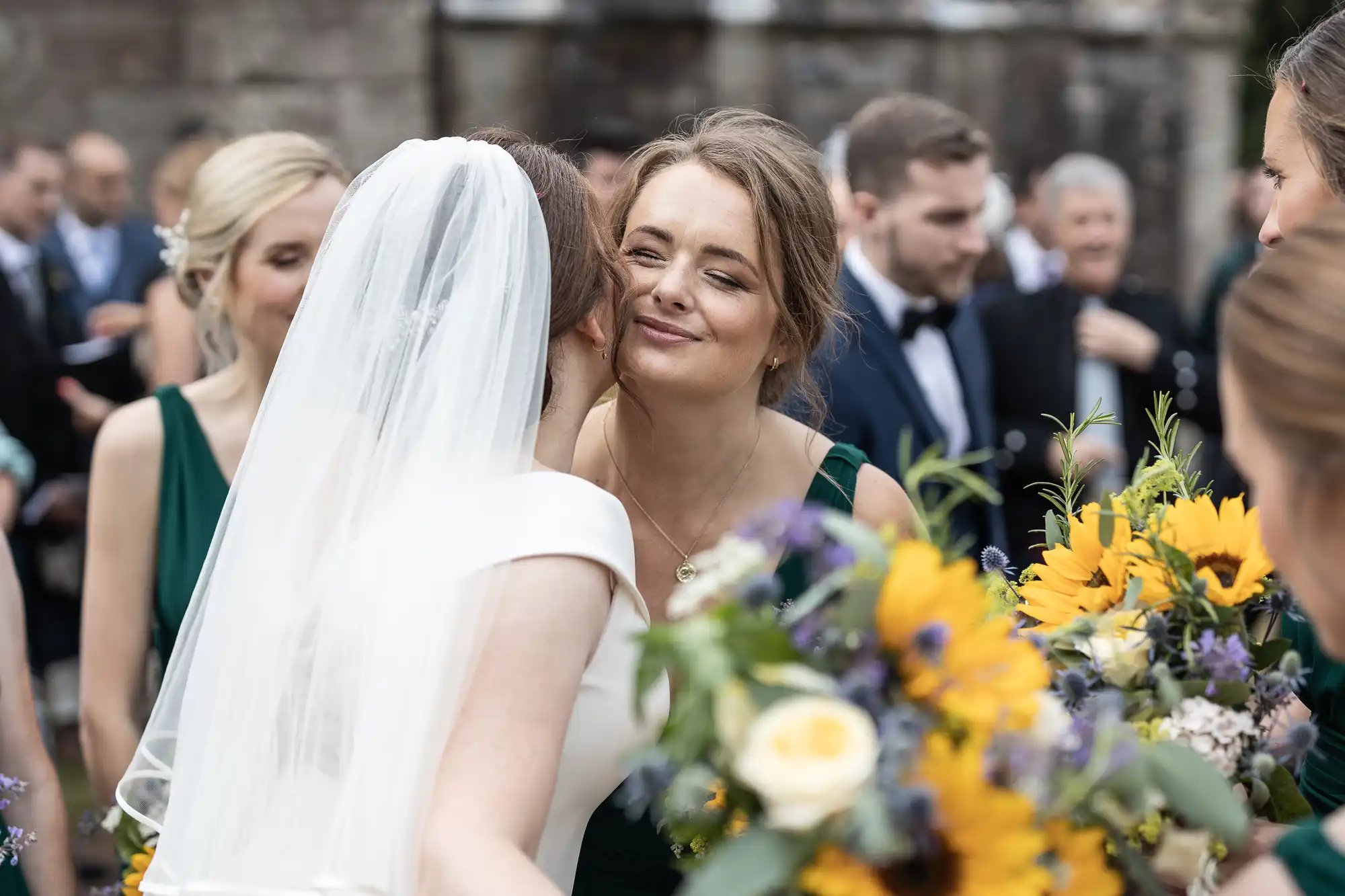 A bride in a white dress and veil embraces a woman holding sunflower-themed bouquets at an outdoor gathering with people in the background.