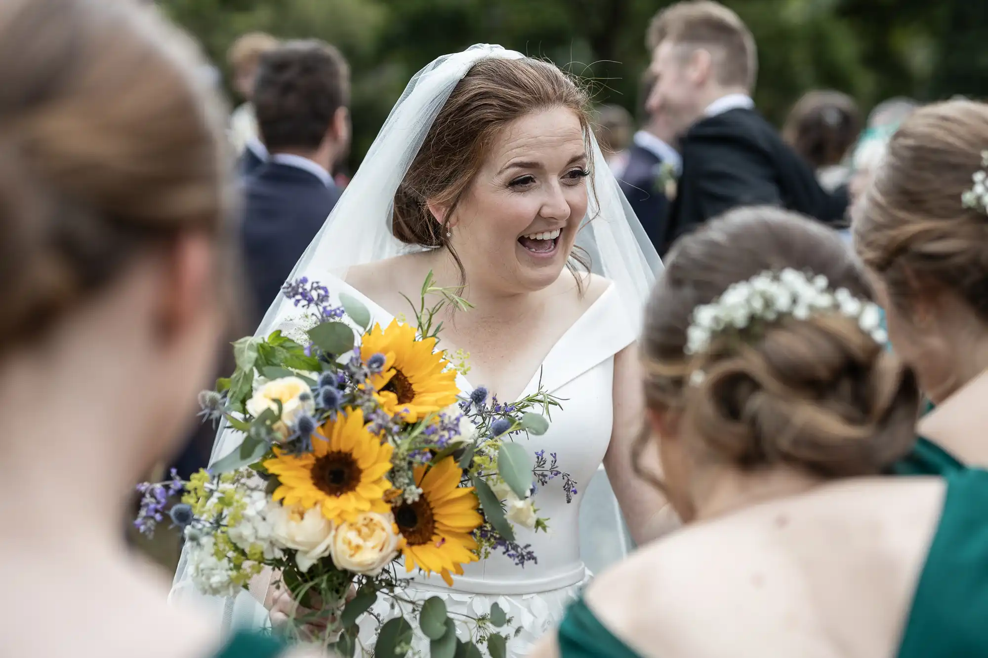 A bride in a white wedding dress, holding a bouquet of sunflowers and other flowers, smiles while interacting with bridesmaids in green dresses.