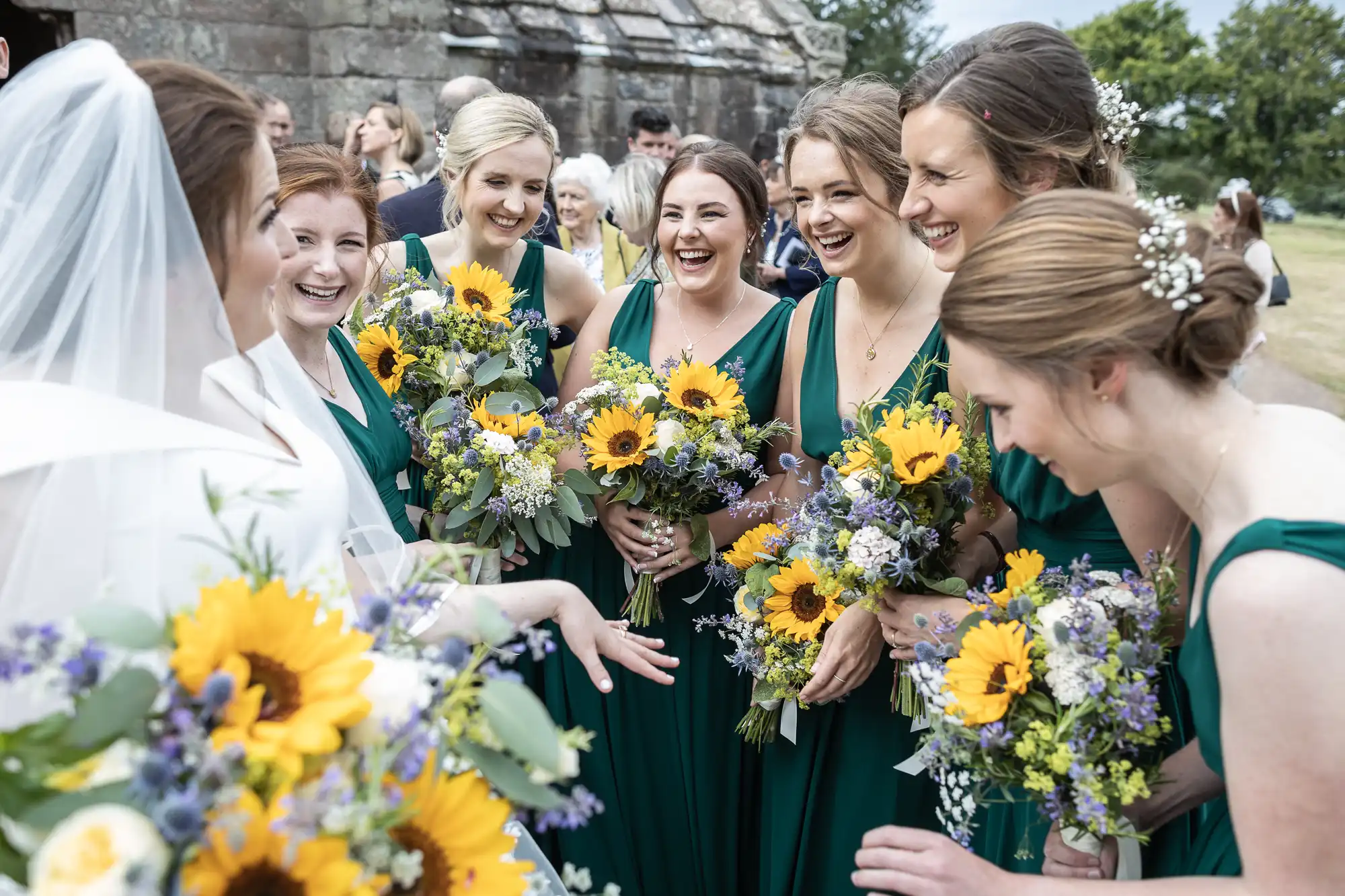 A bride in a white dress is surrounded by six bridesmaids in dark green dresses holding bouquets with sunflowers and other flowers. They are all smiling and standing outdoors.