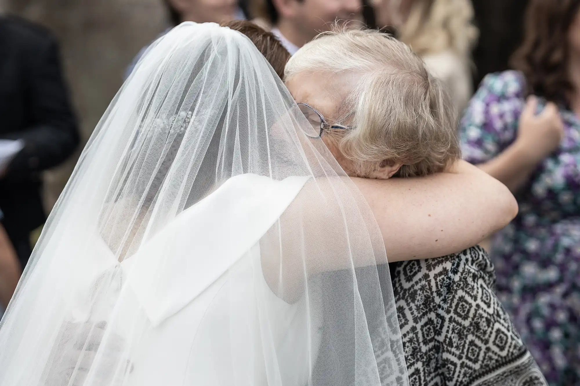 A bride wearing a veil hugs an older woman with glasses during an outdoor event.