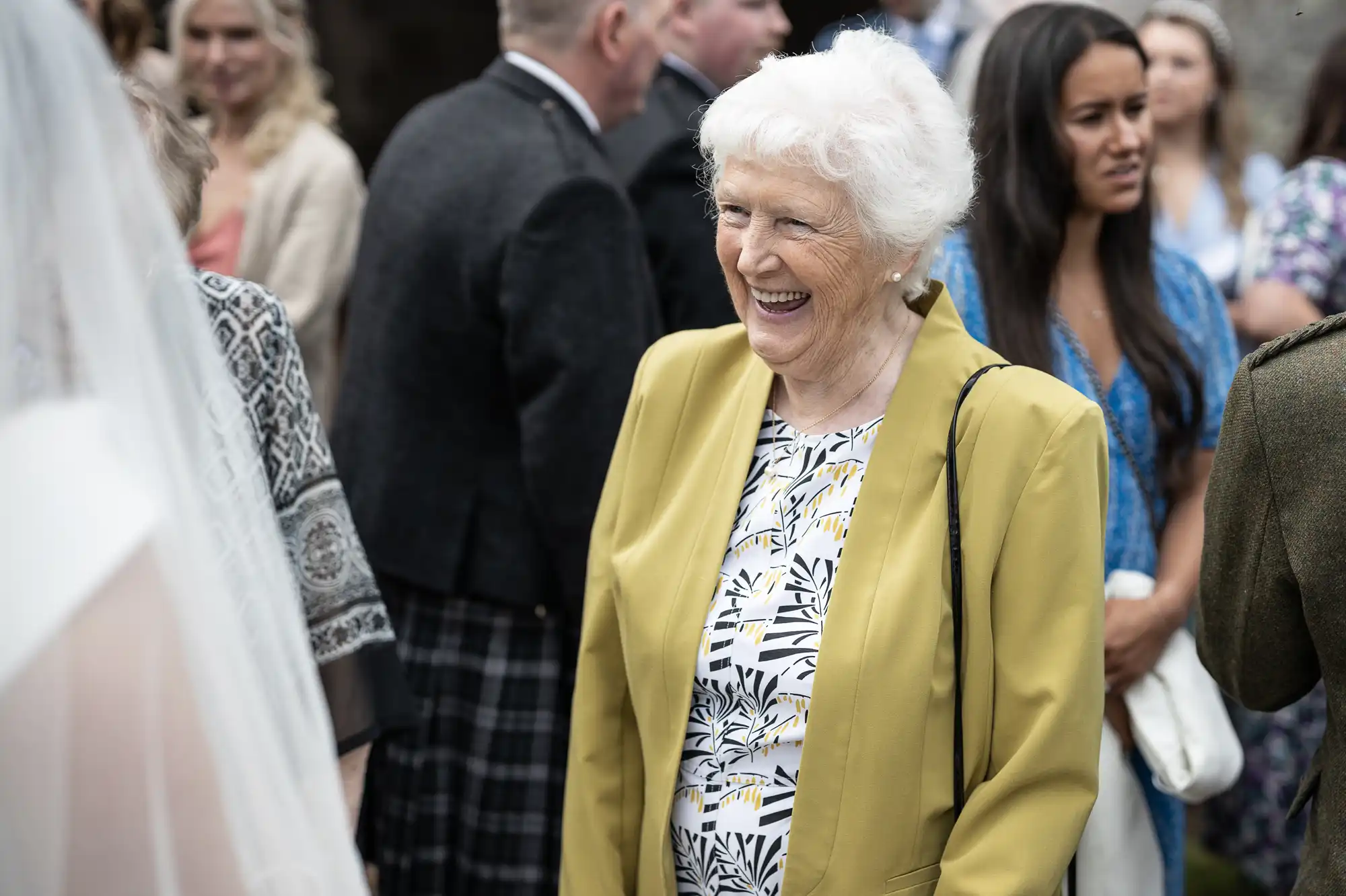 An elderly woman with short white hair, wearing a yellow jacket and patterned blouse, smiles while surrounded by other people at an outdoor event.