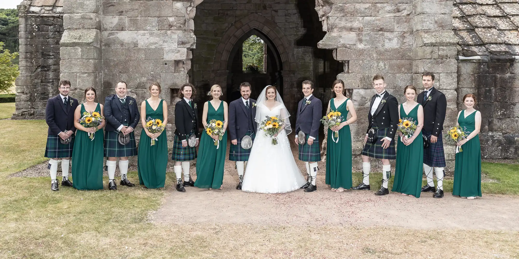 A bride, groom, and their wedding party pose in front of a stone archway. The groomsmen wear kilts, and the bridesmaids wear green dresses with sunflower bouquets.