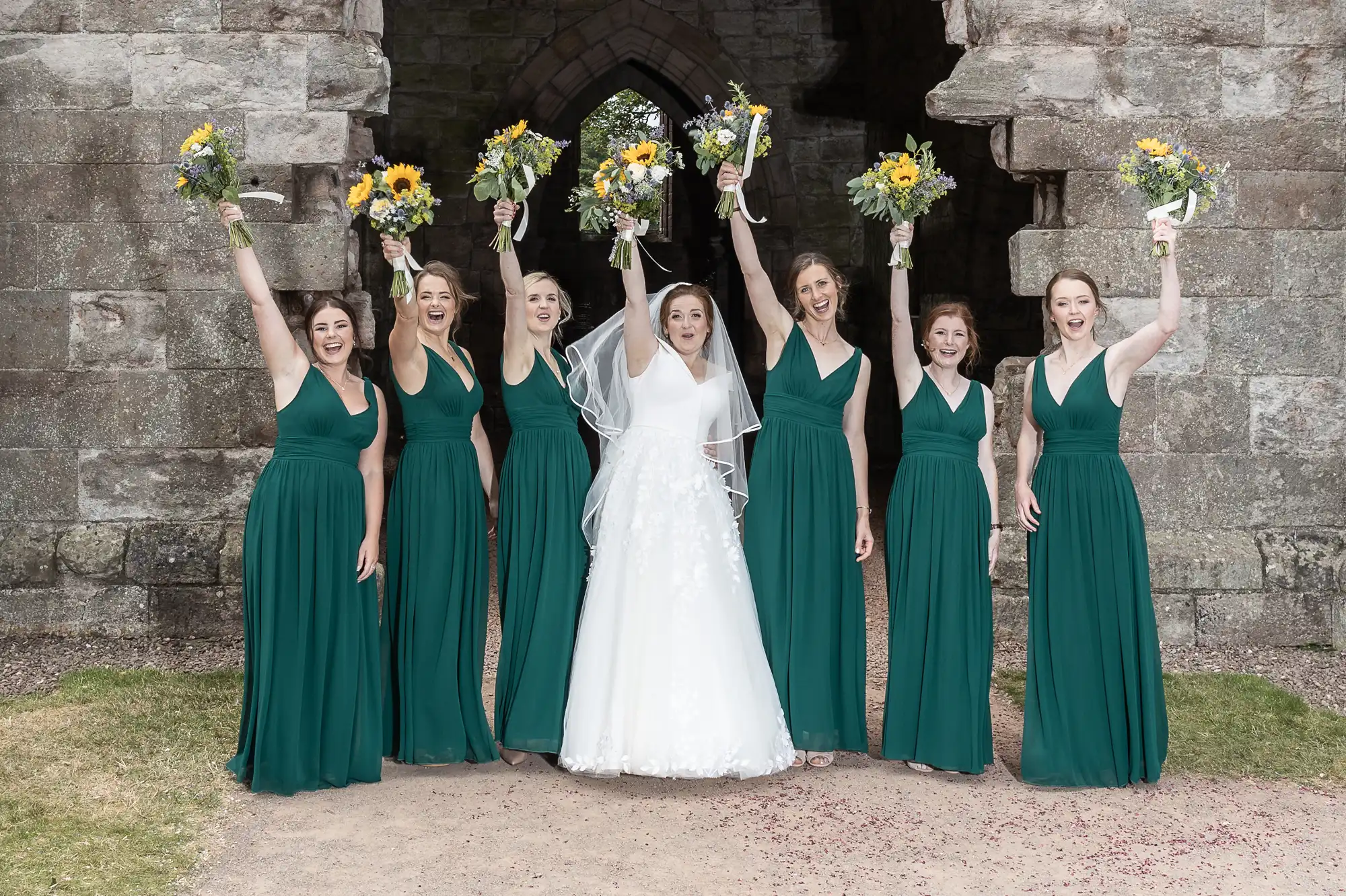 A bride and six bridesmaids in green dresses stand in front of a stone archway, holding bouquets and raising their arms in celebration.