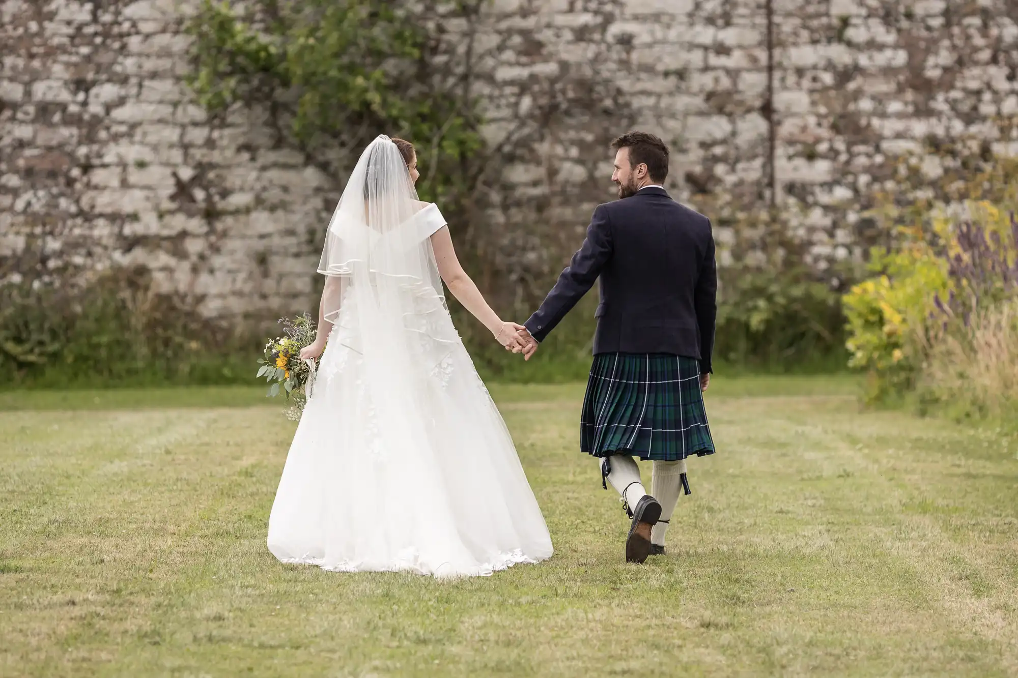 A bride in a white dress and a groom in a kilt walk hand-in-hand on a grassy area with a stone wall in the background.