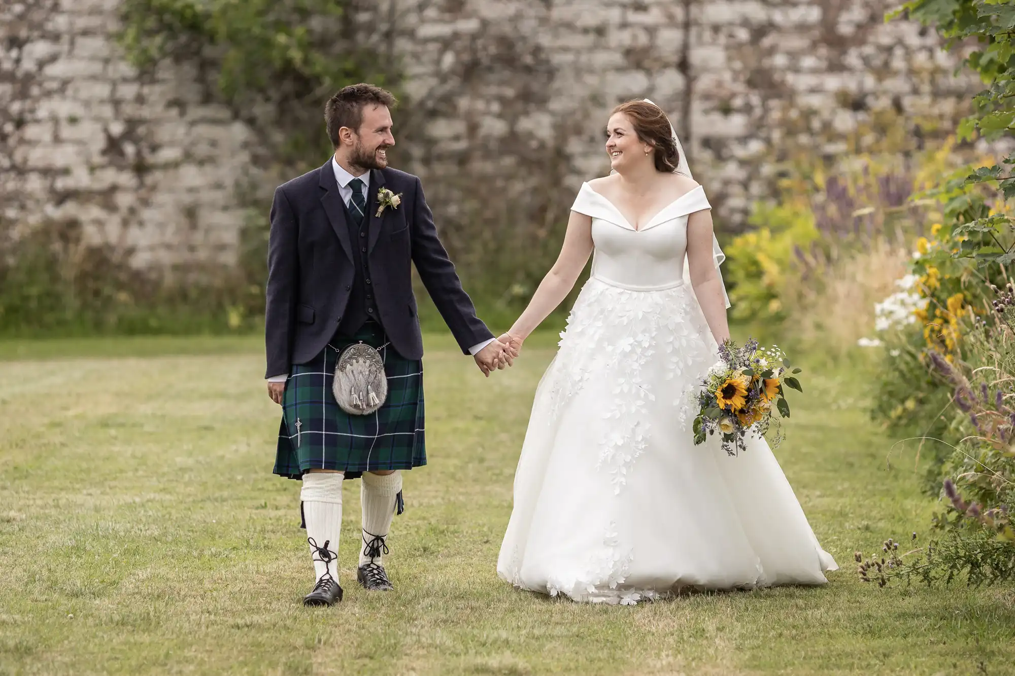 A couple holding hands, walking outdoors on grass; the man is in a kilt, and the woman is in a white wedding dress holding a bouquet of sunflowers.