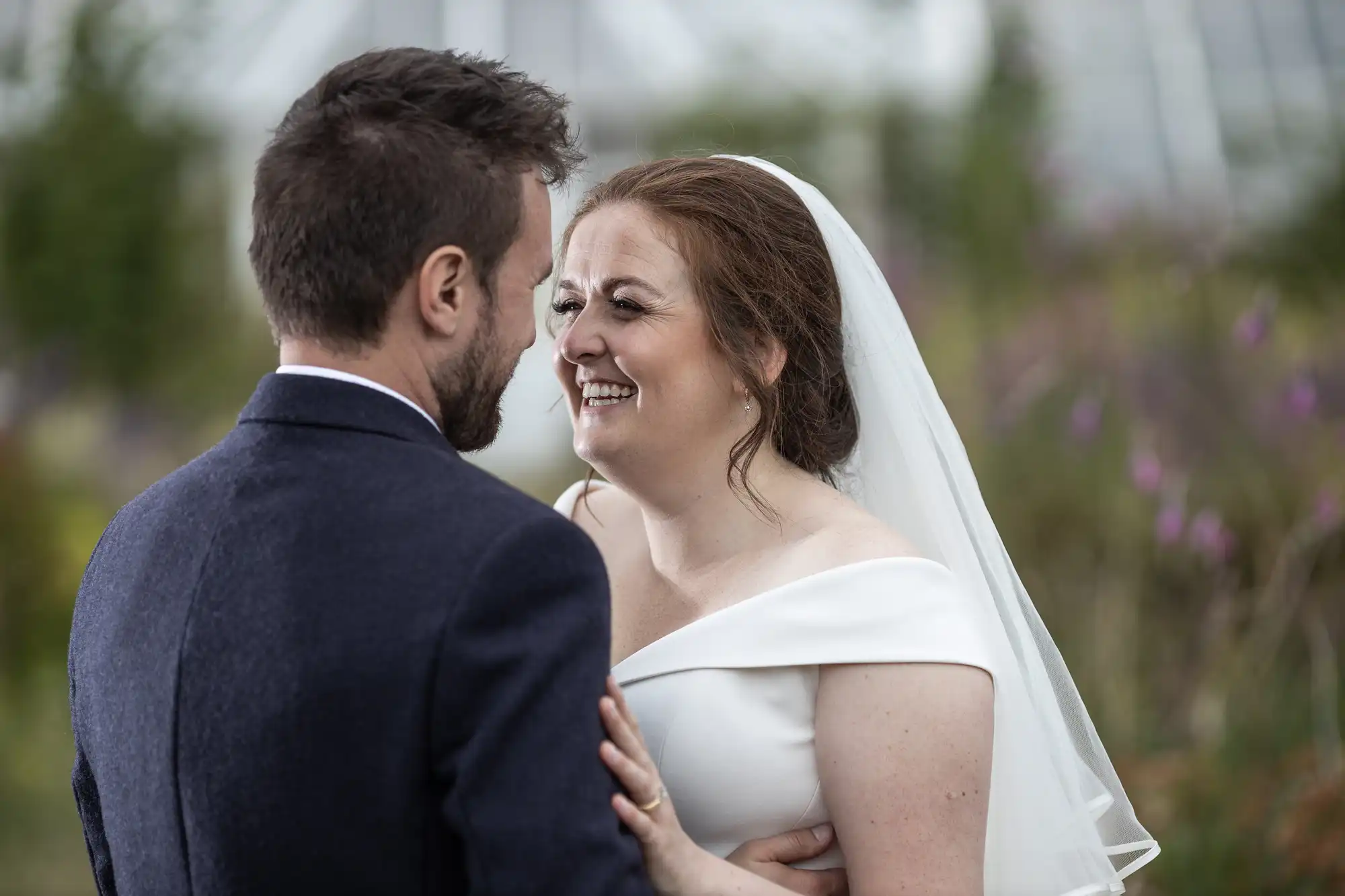 A bride and groom stand facing each other, smiling and holding hands outdoors. The bride wears a white dress and veil, while the groom wears a dark suit.