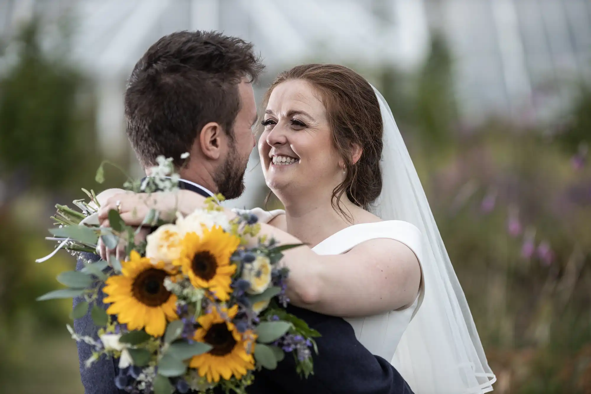 A bride and groom embrace while smiling at each other. The bride holds a bouquet with sunflowers and various greenery.