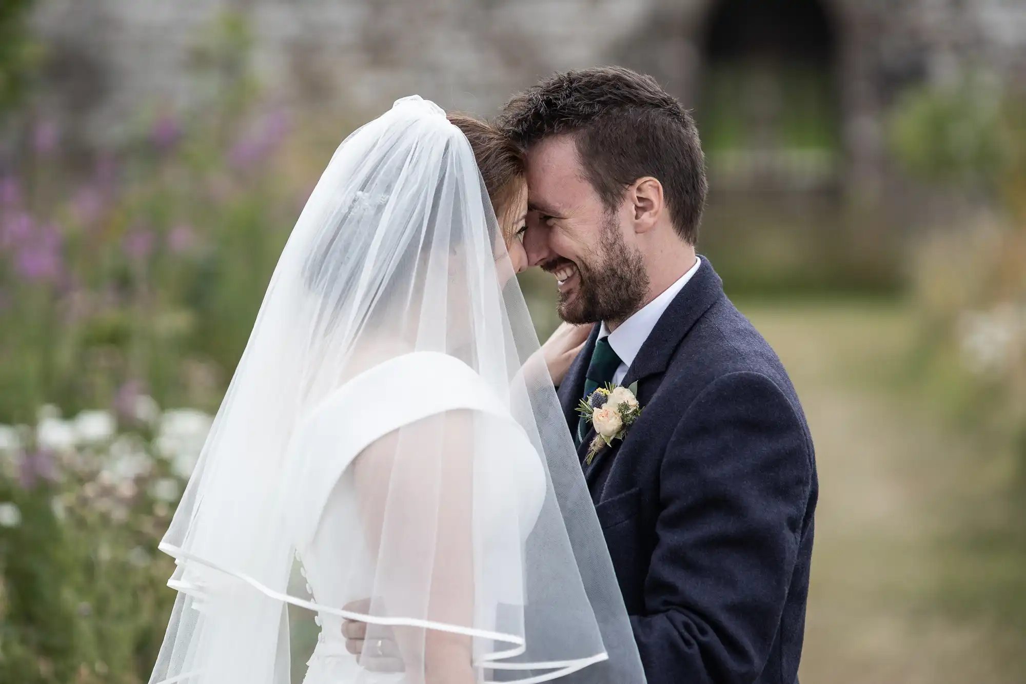 A bride and groom face each other and smile, with the bride wearing a white dress and veil, and the groom in a dark suit, in an outdoor setting with greenery and flowers.