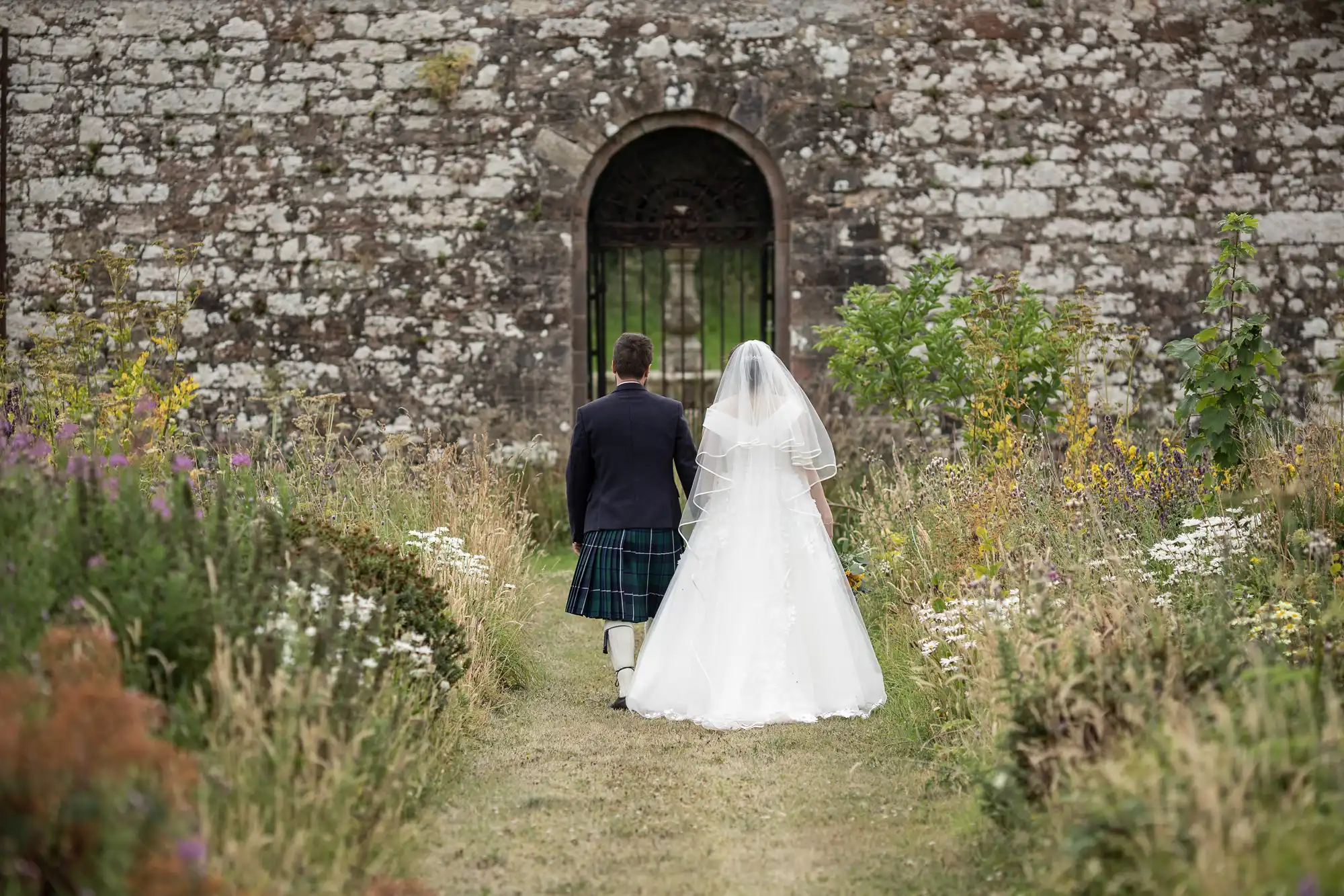 A couple dressed in wedding attire, with the groom in a kilt and the bride in a gown with a veil, walk towards an old stone building through a garden path.