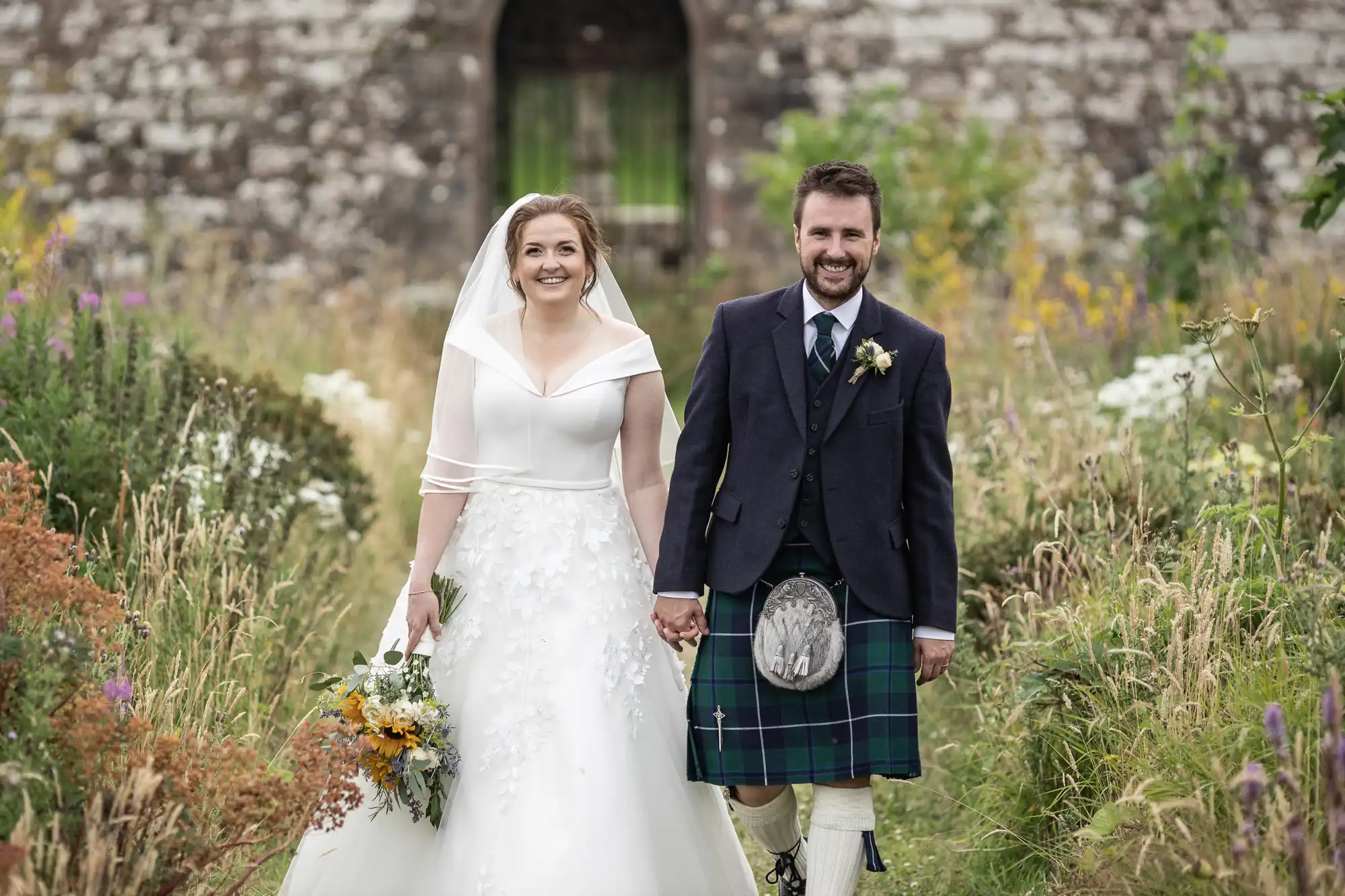 A bride in a white gown and a groom in a kilt walk hand-in-hand down a path in a garden, with a stone building in the background.