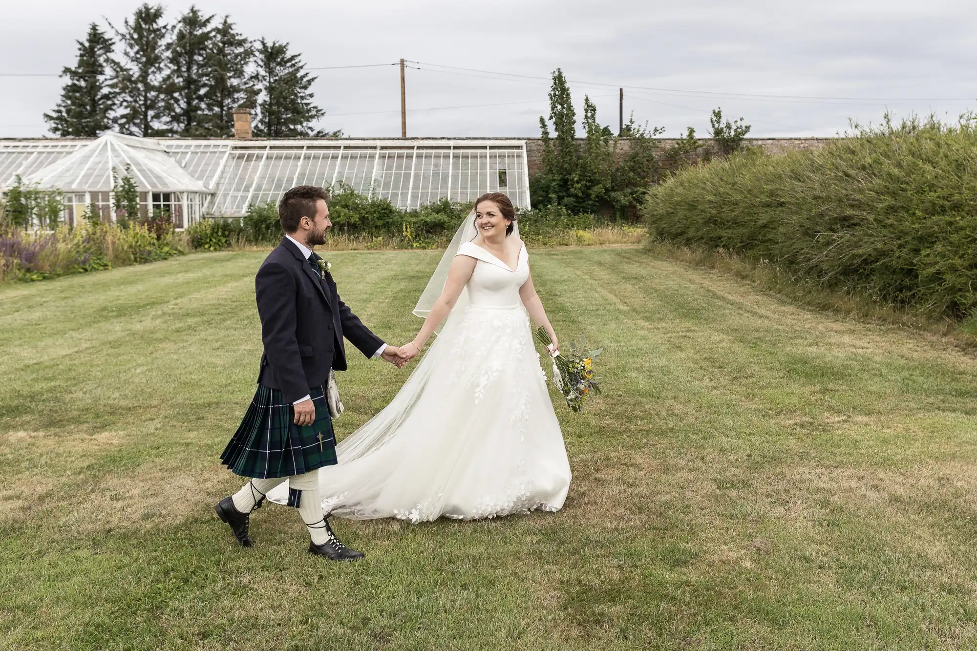 A bride and groom walk hand in hand across a grassy area, with the groom wearing a kilt and the bride in a white gown. Greenhouses and trees are visible in the background.