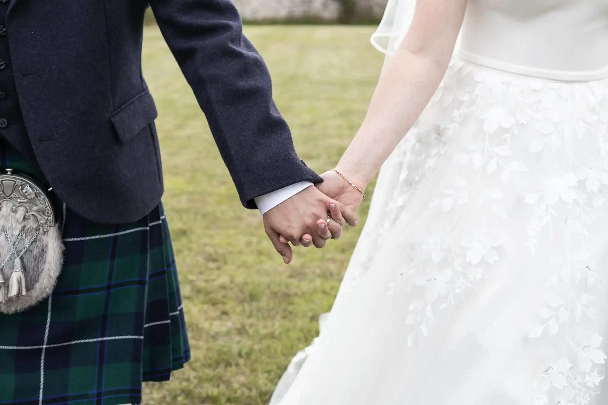 A couple dressed in formal attire, with the person on the left wearing a kilt and the person on the right wearing a white dress, hold hands while standing outside on grass.