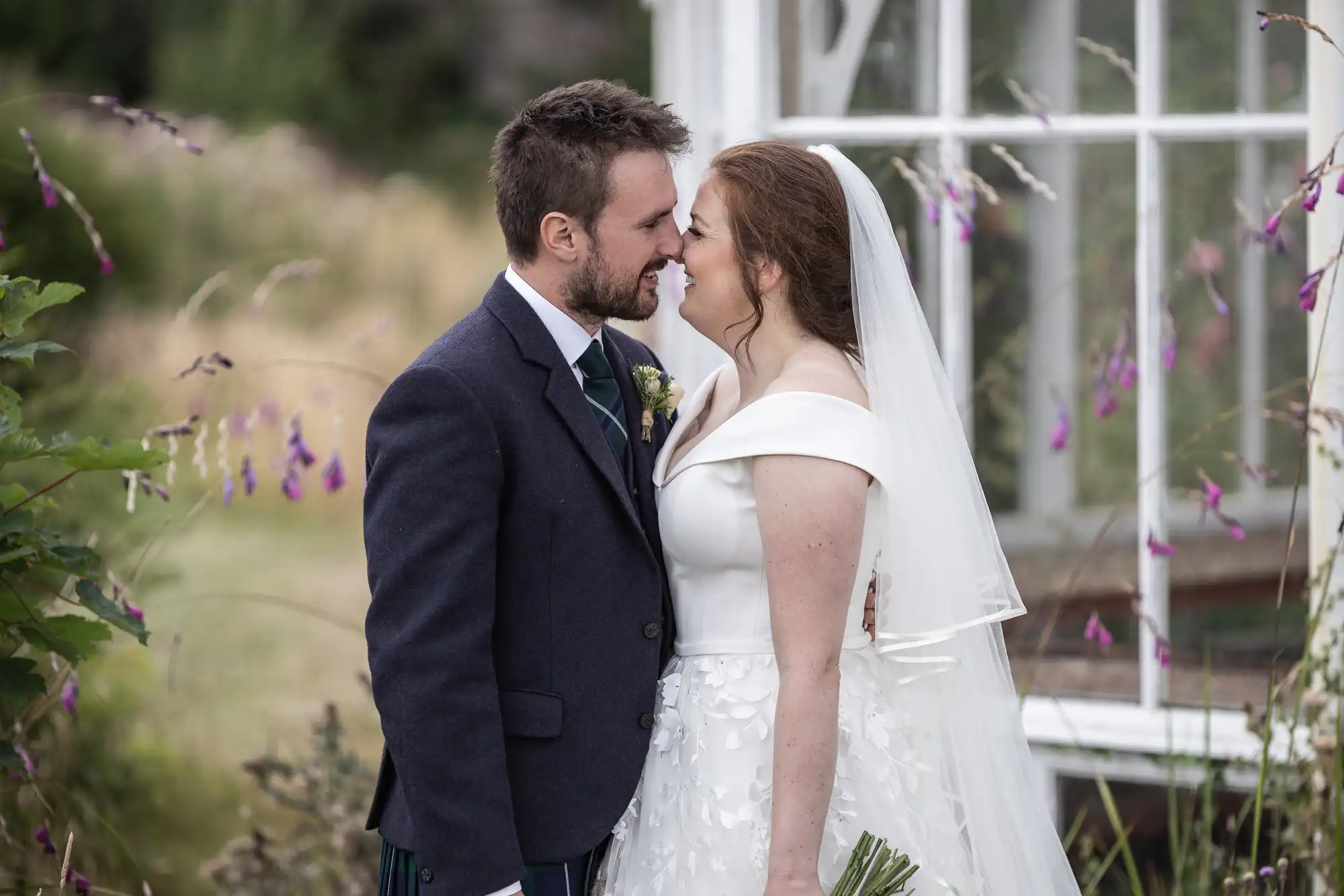 A bride and groom stand close, touching foreheads, in front of a glass-windowed building with greenery and purple flowers surrounding them.