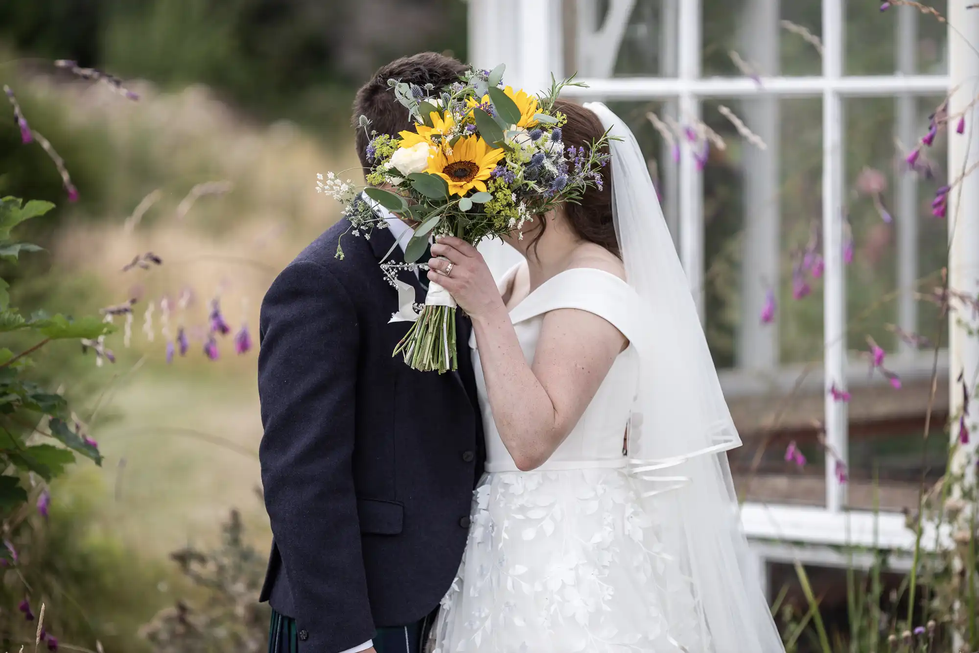 A bride and groom standing outside share a kiss, partially hidden behind a bouquet of flowers. The bride is wearing a white dress and veil, while the groom is in a dark suit.