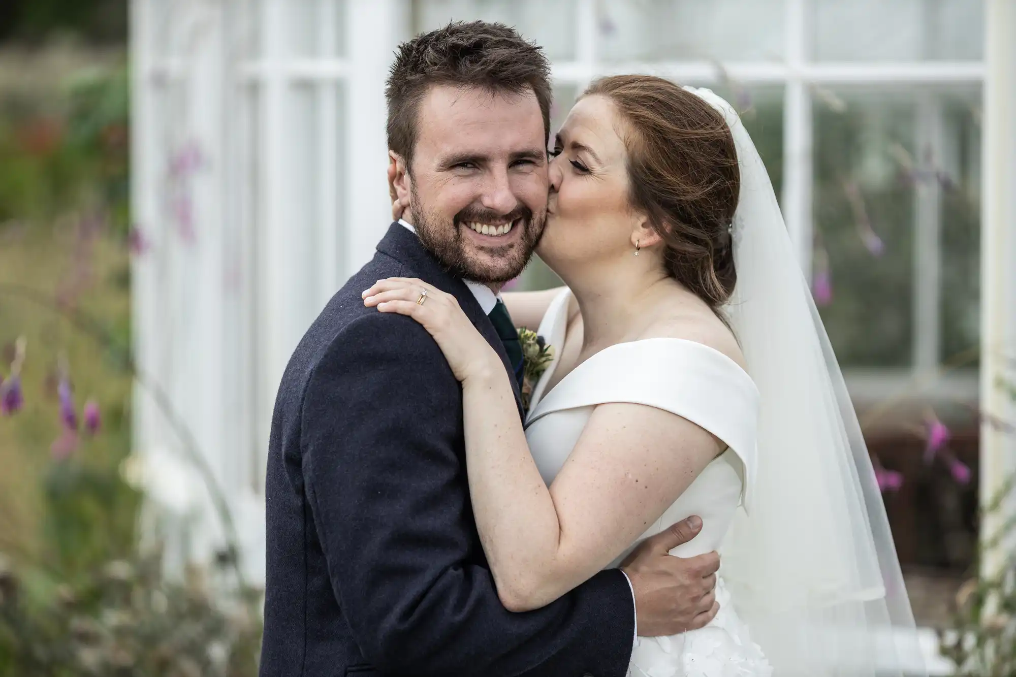 A bride, wearing a white dress and veil, kisses the cheek of a smiling groom dressed in a dark suit, as they embrace in front of a window.