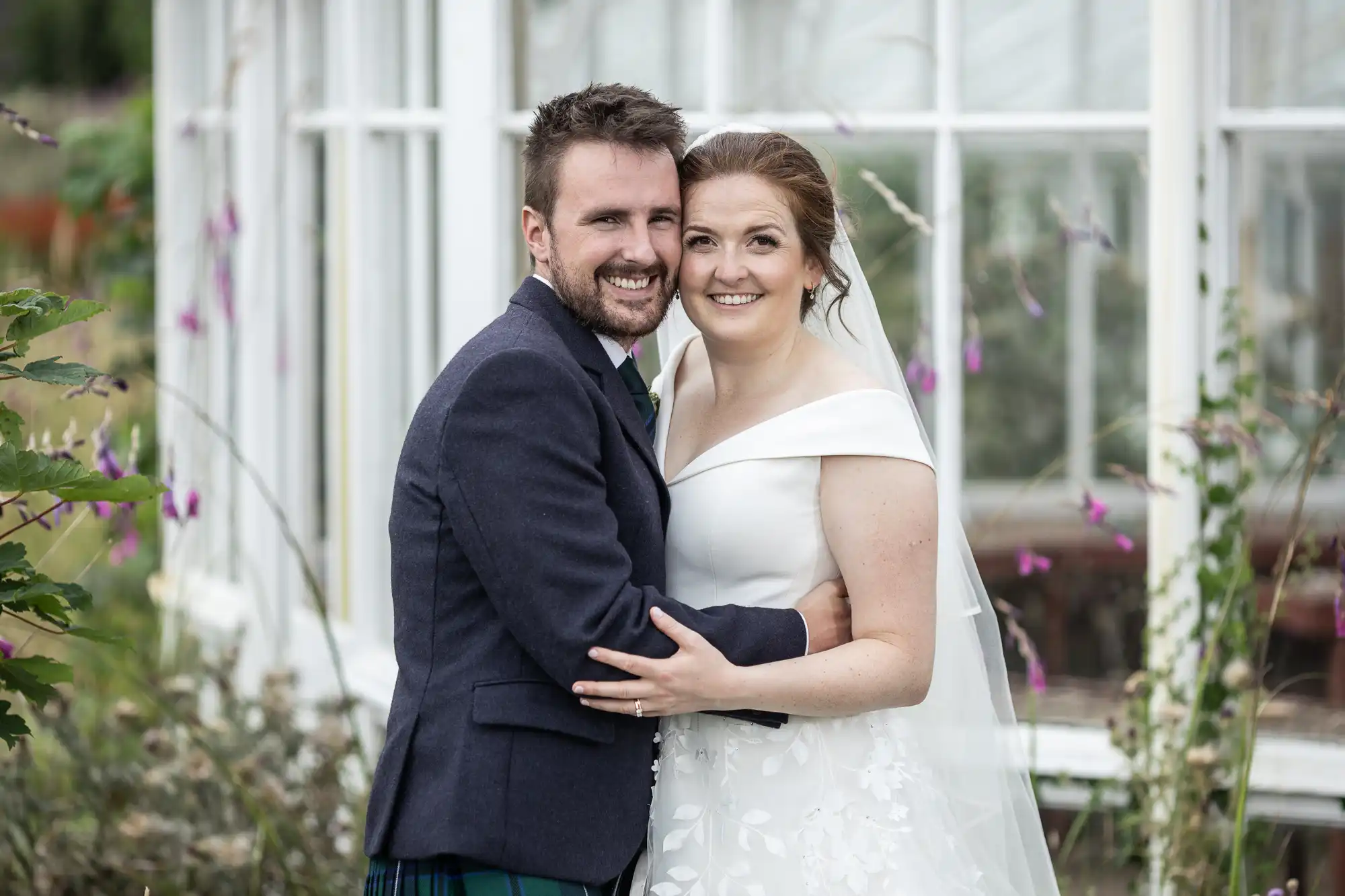 A couple in formal attire smiles and embraces in front of a large window, with plants around them.