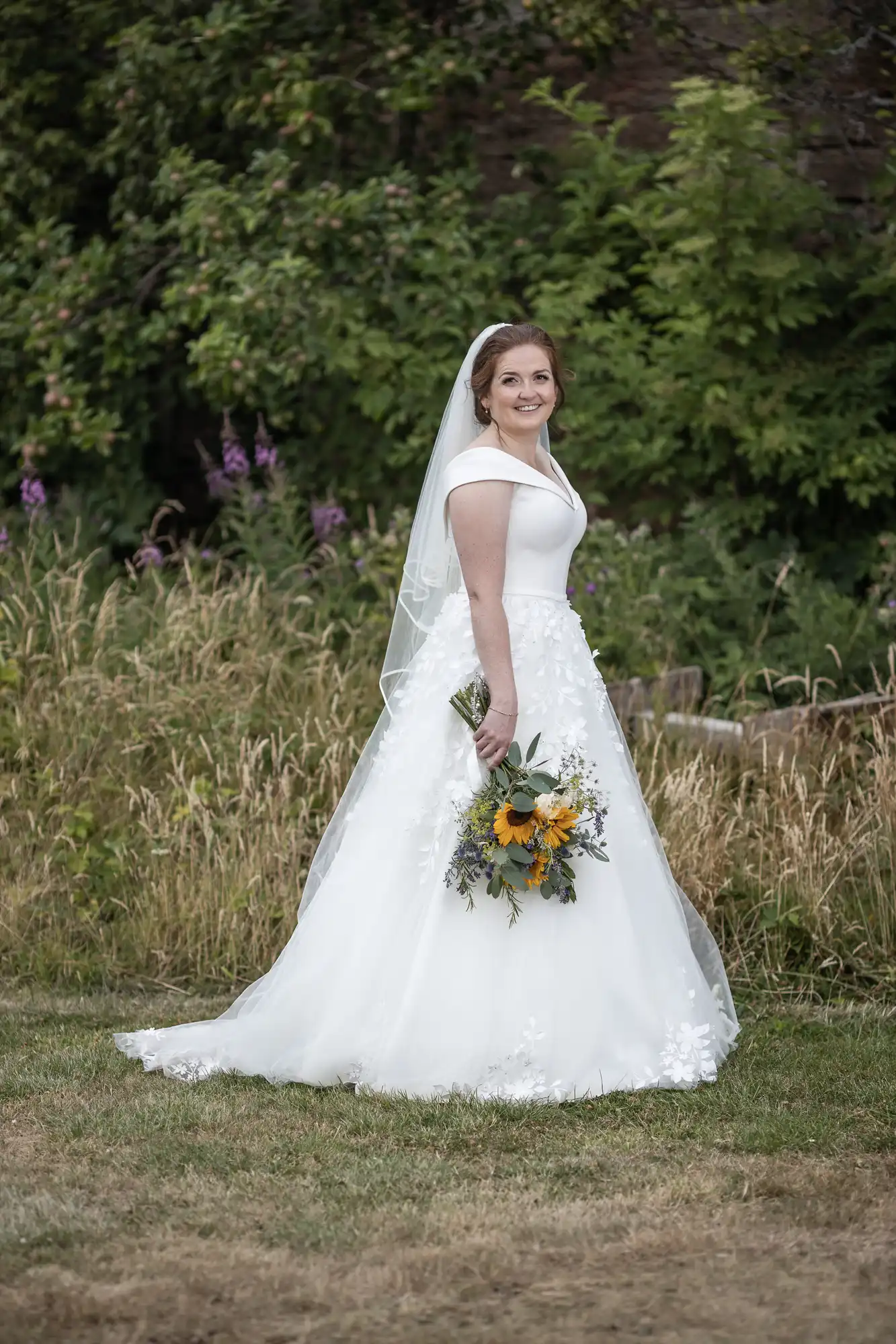 A bride in a white wedding dress and veil stands outdoors on grass, holding a bouquet of flowers, with greenery in the background.