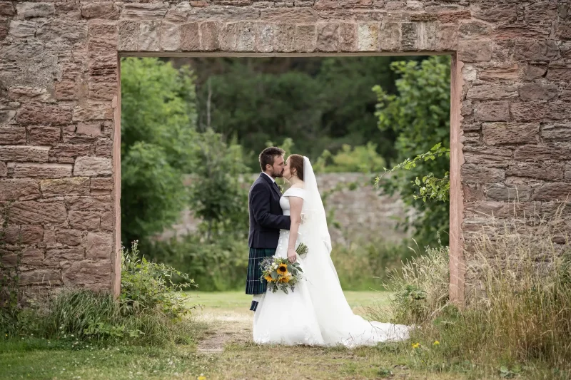 wedding at Dunglass Estate - A bride and groom stand kissing under a stone archway, with greenery in the background. The bride is wearing a white gown and veil, holding a bouquet of sunflowers, while the groom wears a kilt.