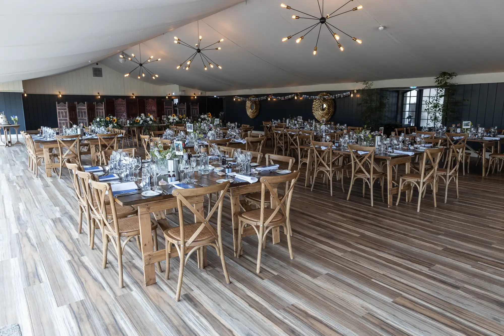 A large, elegantly decorated reception hall with wooden tables and chairs, set for a formal event. The tables are adorned with glassware, cutlery, floral centerpieces, and napkins. Multiple chandeliers hang from the ceiling.