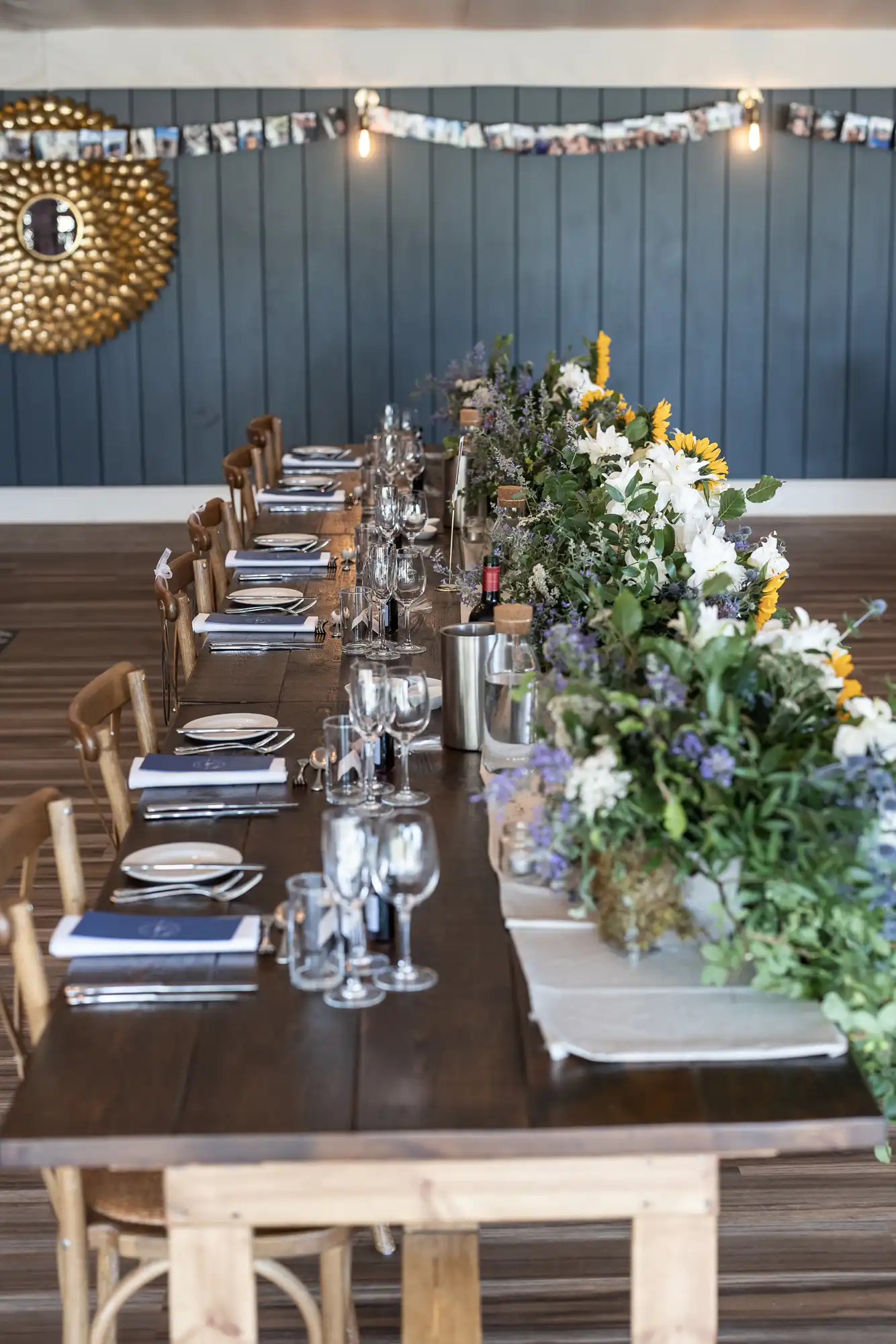 A long wooden table is set for a meal with plates, glasses, and cutlery. A floral centerpiece runs down the middle. Polaroid photos are strung up in the background with lights above them.