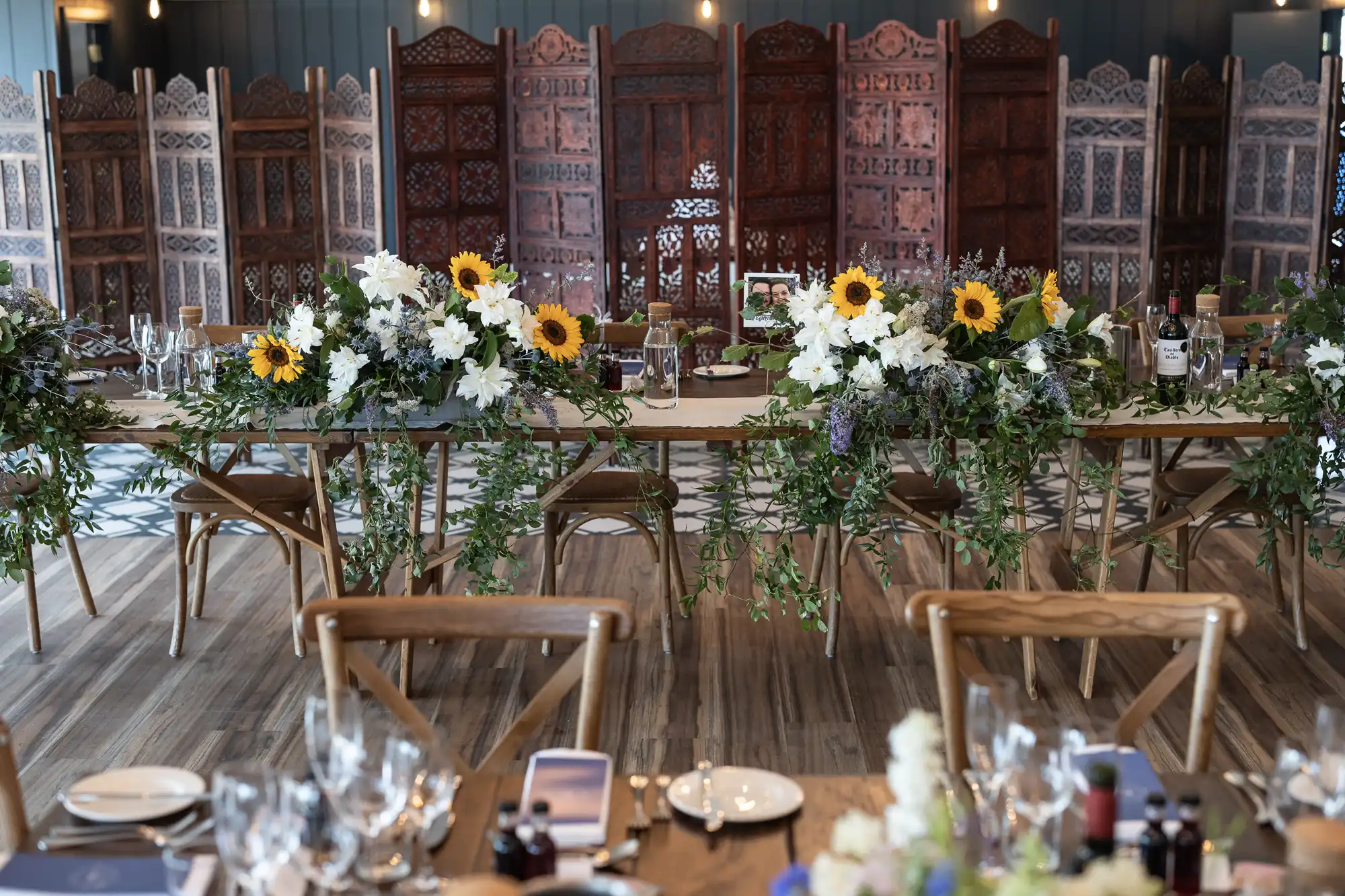 A dining setup with wooden chairs, tables adorned with floral arrangements including sunflowers and white lilies, and decorative wooden panels in the background.