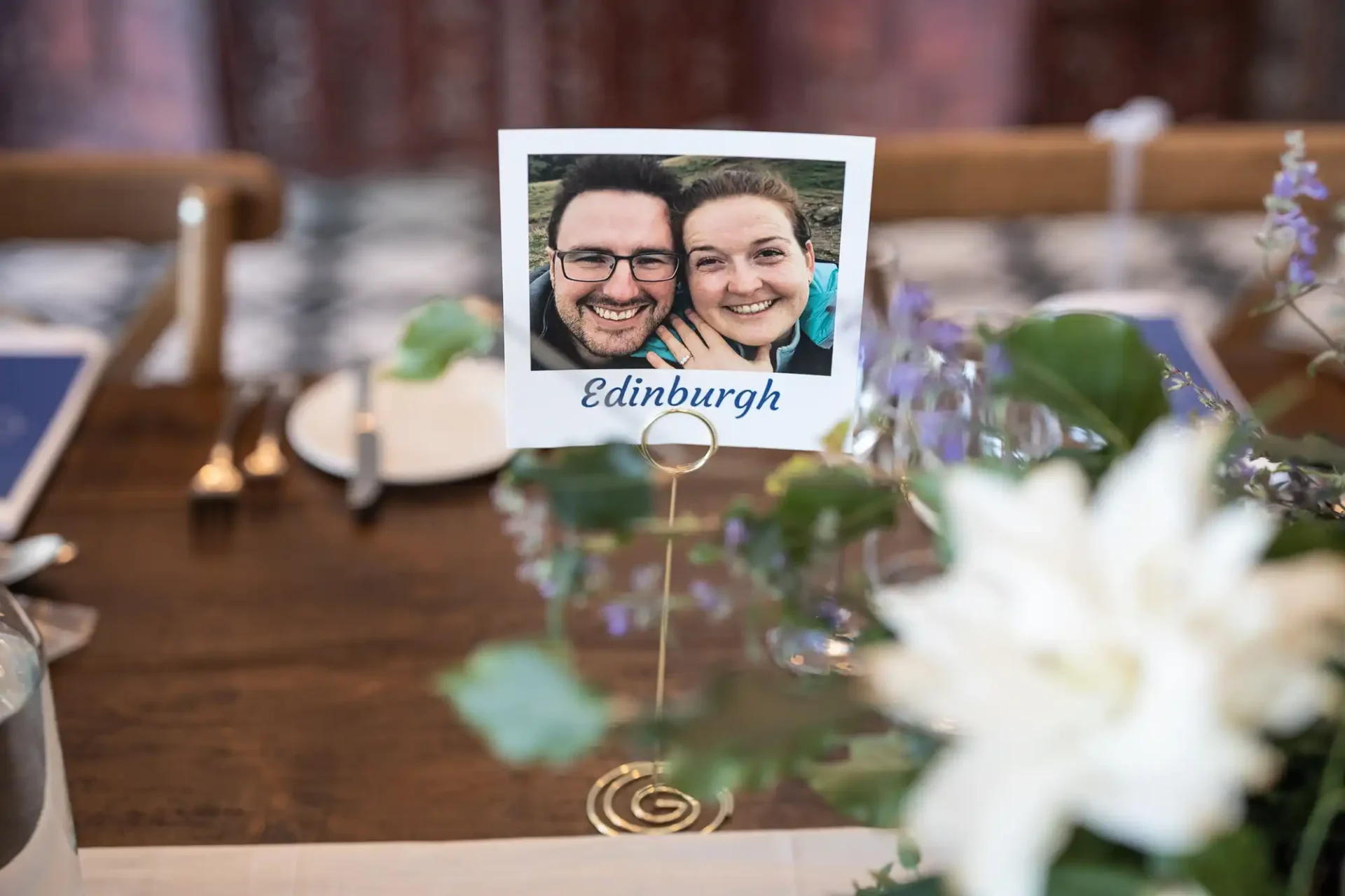 Close-up of a table center display featuring a photo of a smiling couple labeled "Edinburgh." The background shows cutlery, plates, and blurred greenery with a white flower in the foreground.